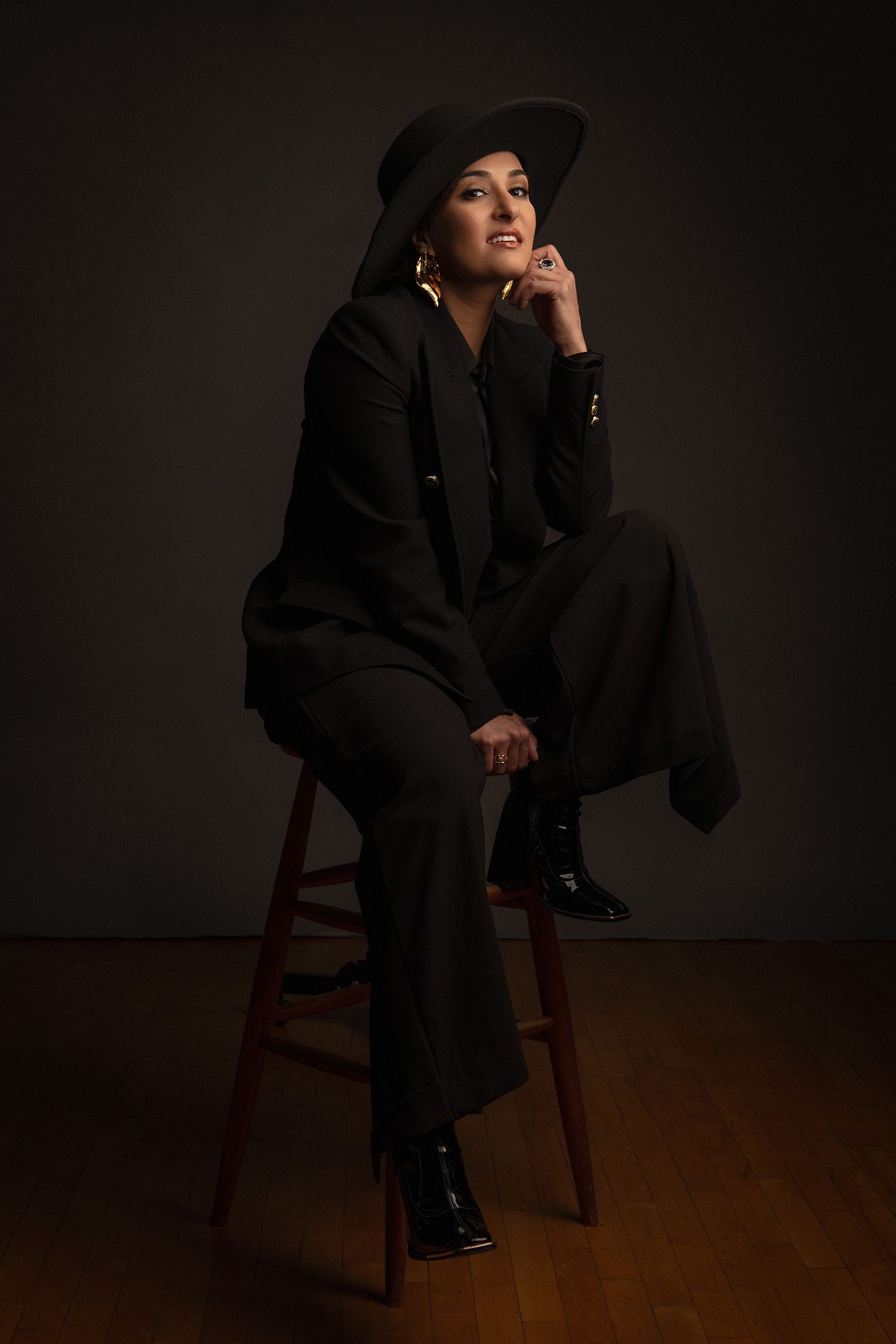 persian pop singer Rana Mansour in black outfit sits thoughtfully on wooden chair against dark background.