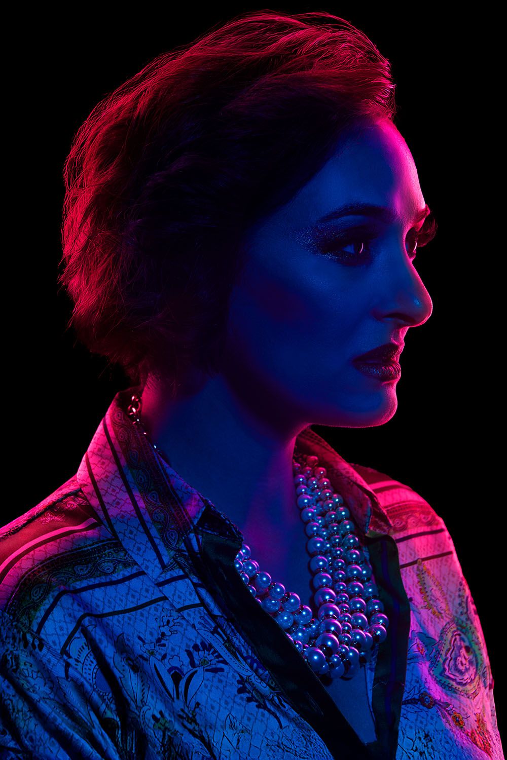 Elegant woman in profile view illuminated by blue and red neon lights wearing a pearl necklace.