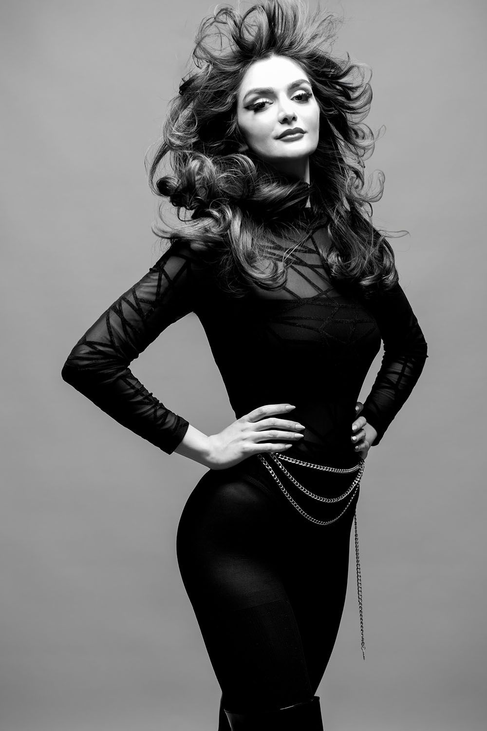 Black and white photo of a glamorous woman in a black dress with chain belt, striking pose with windswept hair.