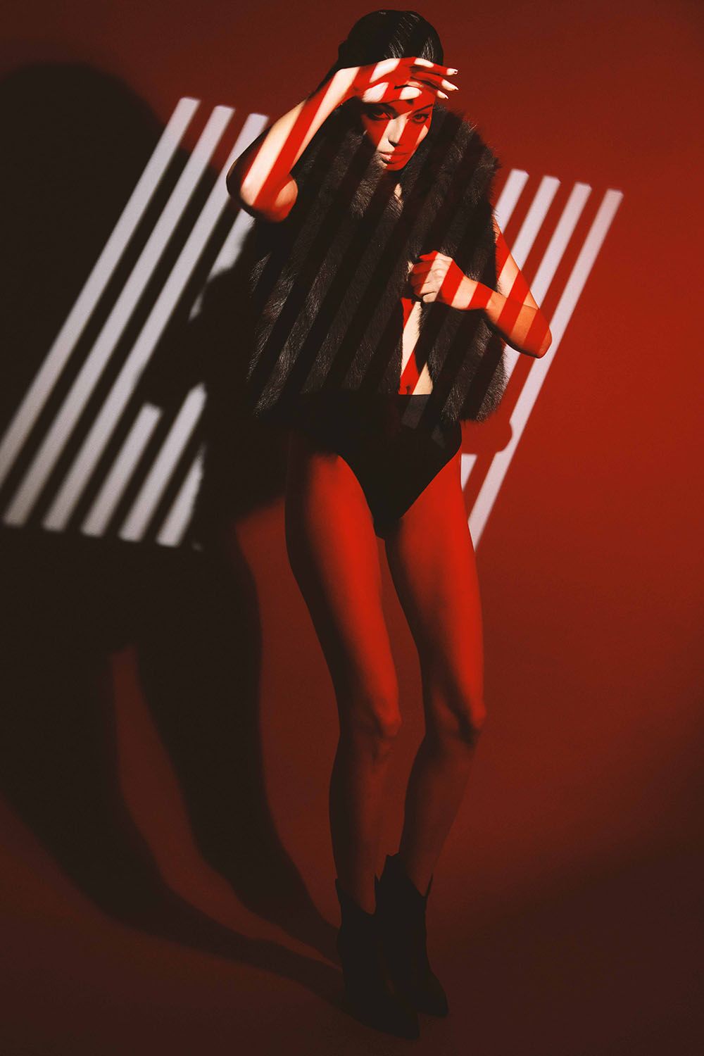 Woman in fur vest and bodysuit poses dramatically under striped red light against a bold red background.