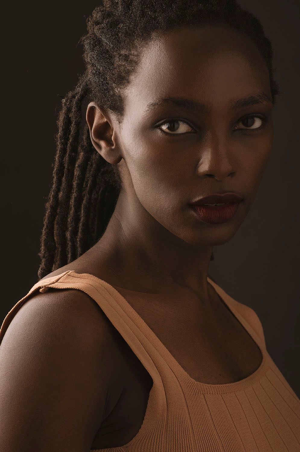 Young Black woman with locs wearing beige tank top, intense gaze in moody studio portrait.
