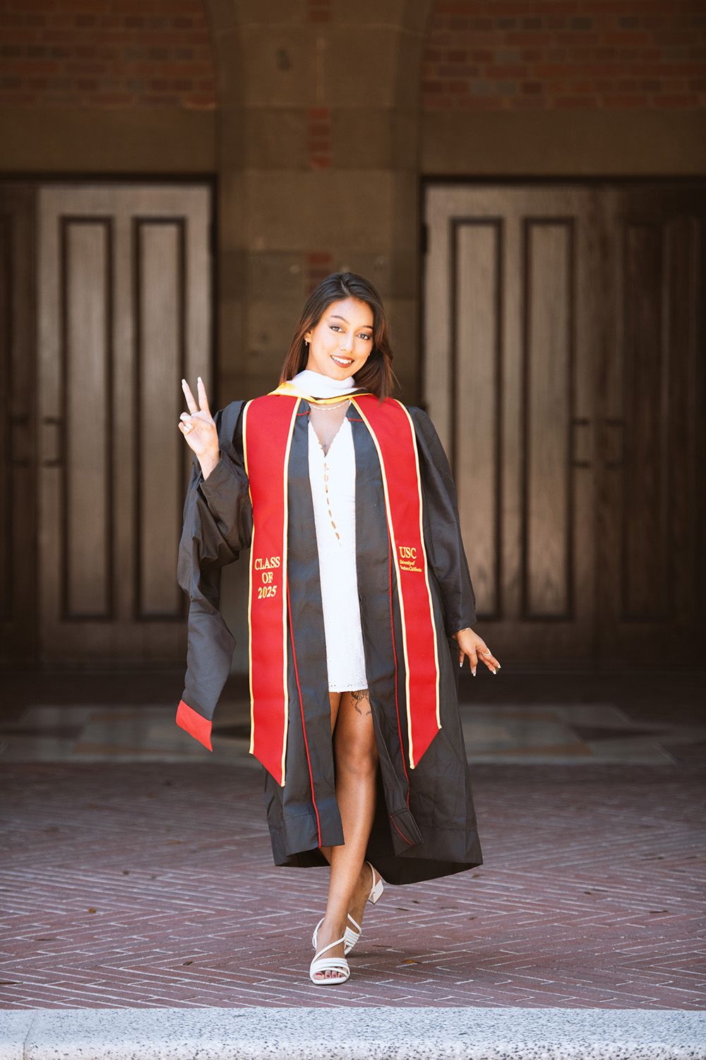 Young woman in black graduation gown with red USC stole posing with peace sign outdoors.