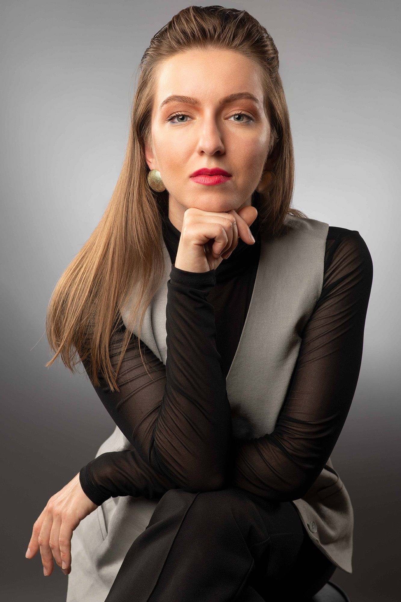 Elegant woman in black sheer top and gray vest poses thoughtfully with hand on chin against gray background.