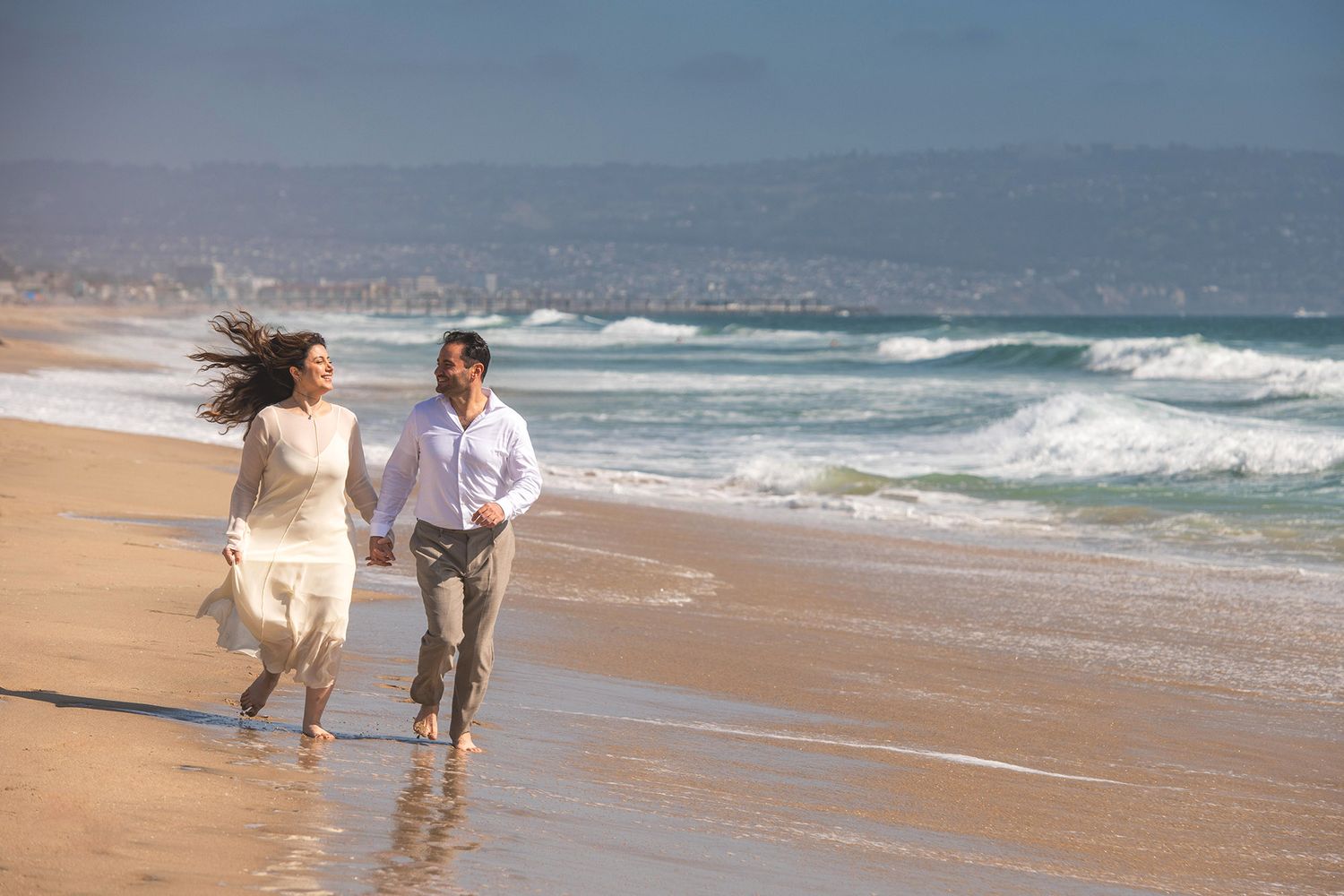 Two people walk along a sandy beach at sunset with waves rolling in from the ocean.