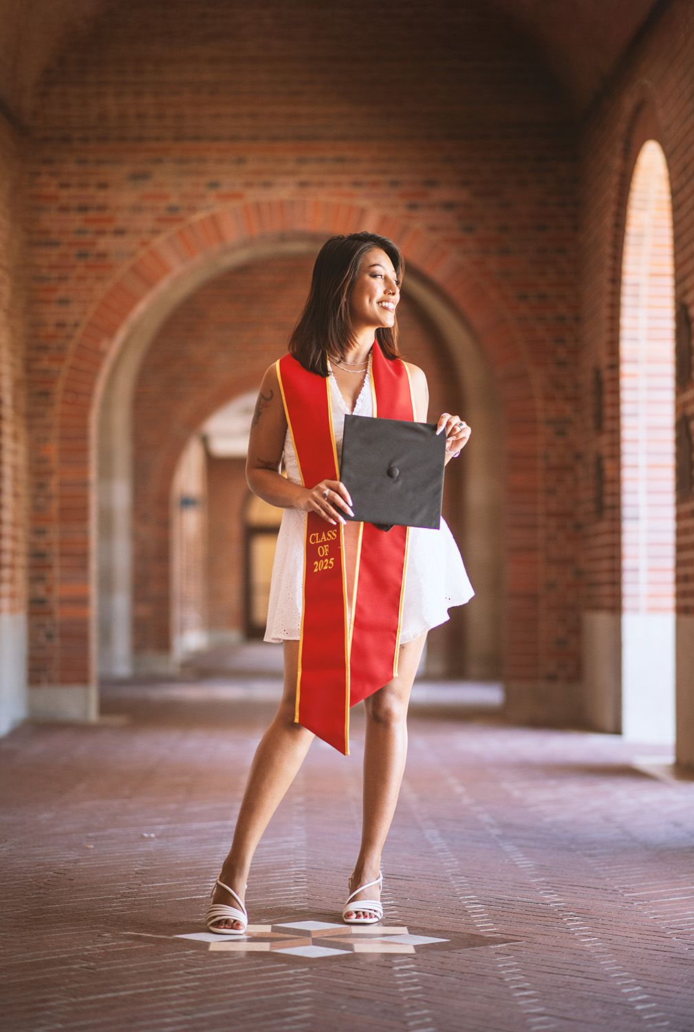 A smiling graduate poses in a brick archway hallway wearing a red sash and holding a cap, Class of 2021.