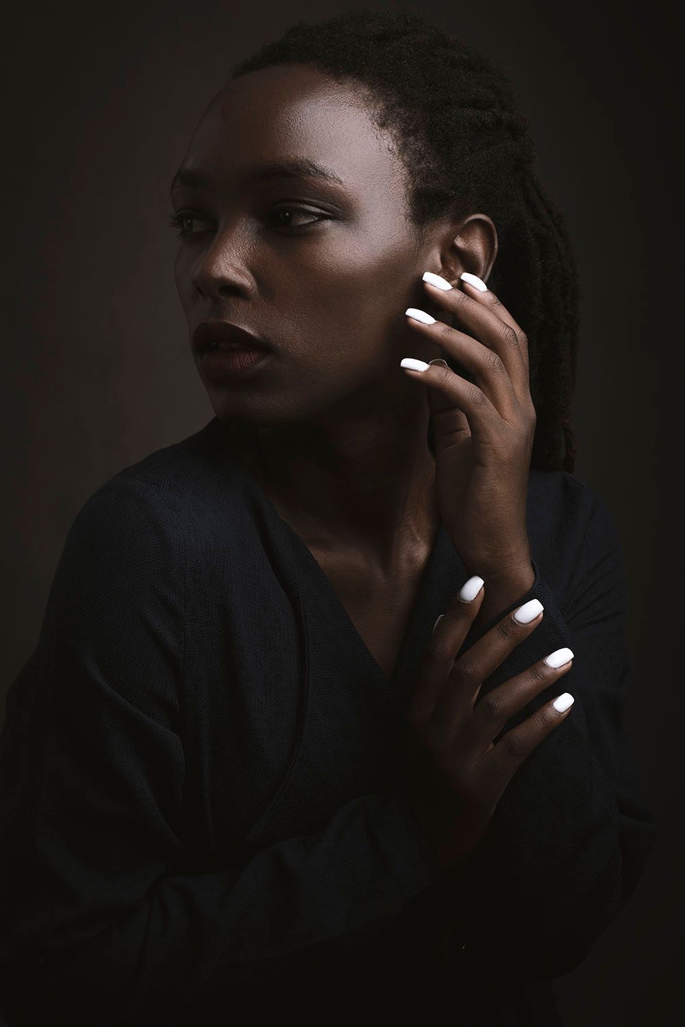 A young Black woman with braids touches her ear, wearing a dark top, with white nails, against a moody dark background.