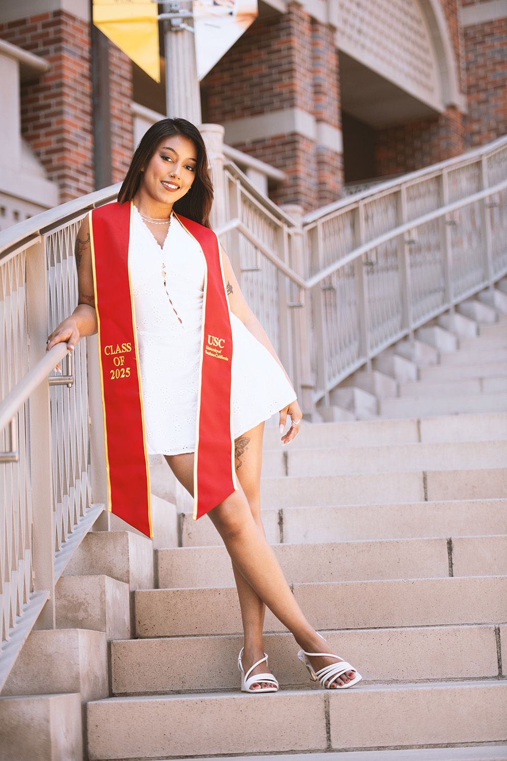Graduate in white dress with red USC Class of 2022 stole posing on outdoor staircase.