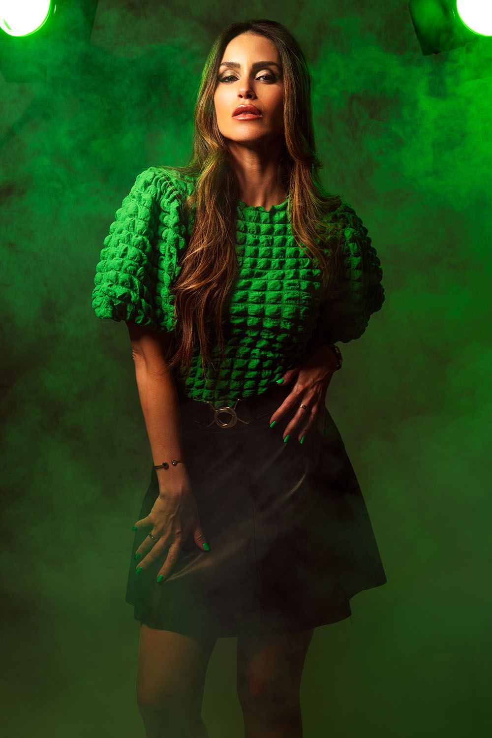 Woman in green textured puff-sleeve top and black mini skirt posing dramatically in green smoky studio lighting.