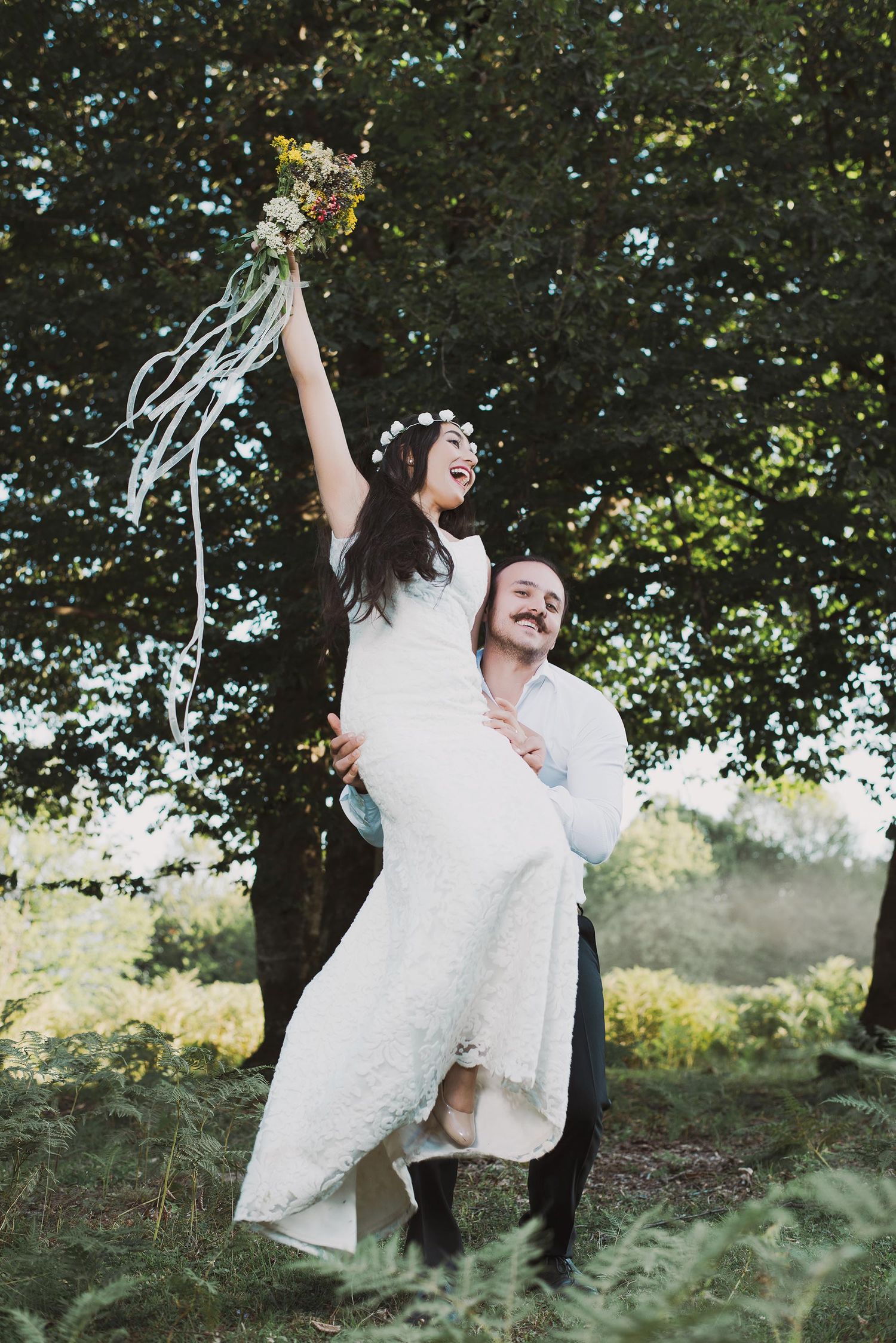 A couple in wedding attire dances joyfully in a natural outdoor setting with trees and greenery in the background.