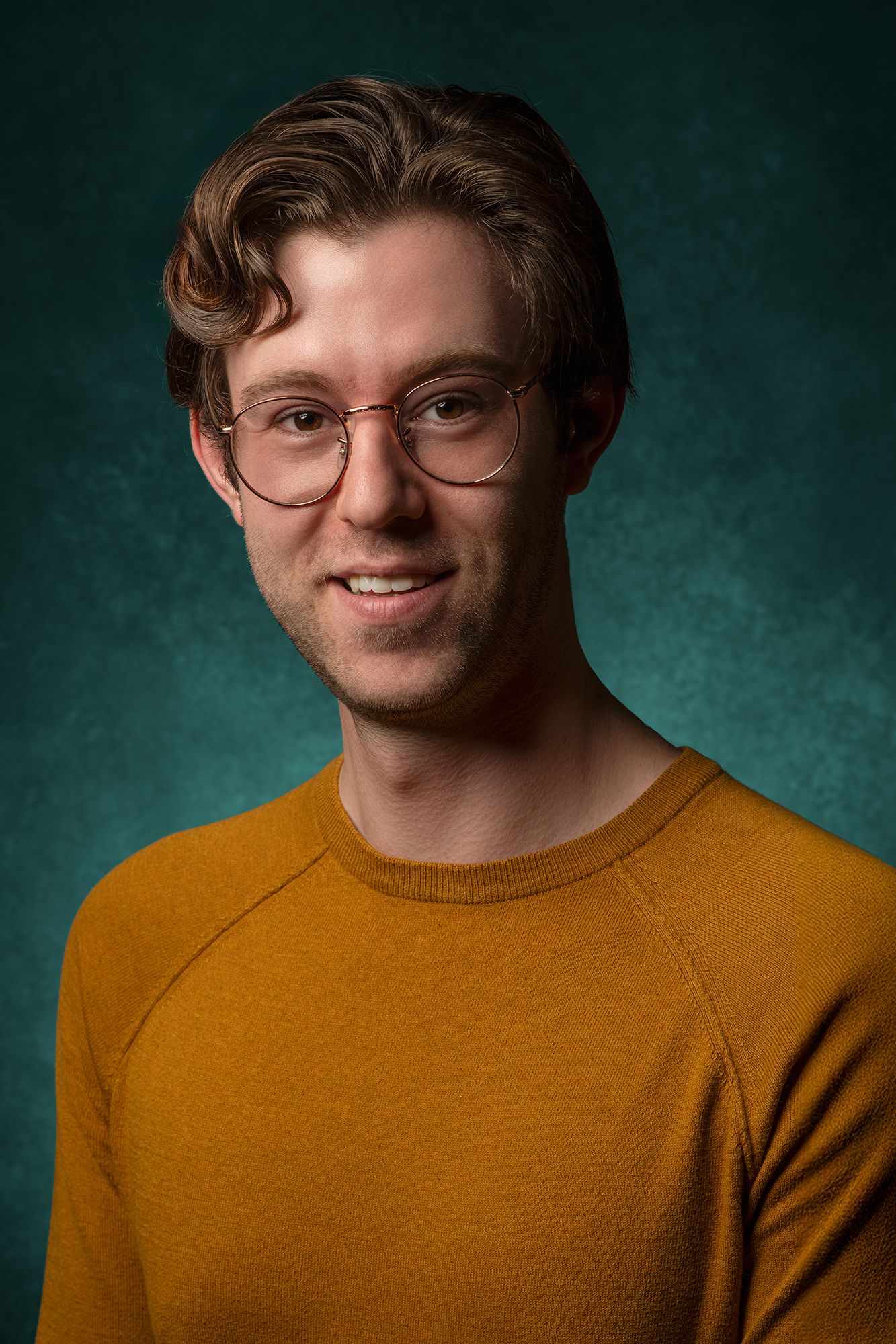 Casting headshot of person in orange sweater and glasses smiling against green background taken in west Los Angeles.