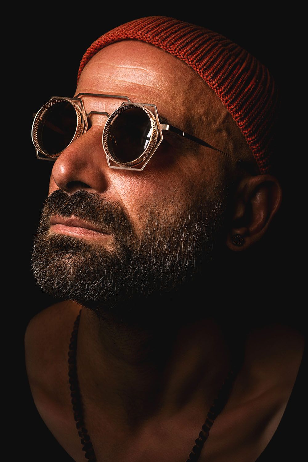 Bearded man wearing round steampunk goggles and red knit beanie in a dark dramatic portrait.
