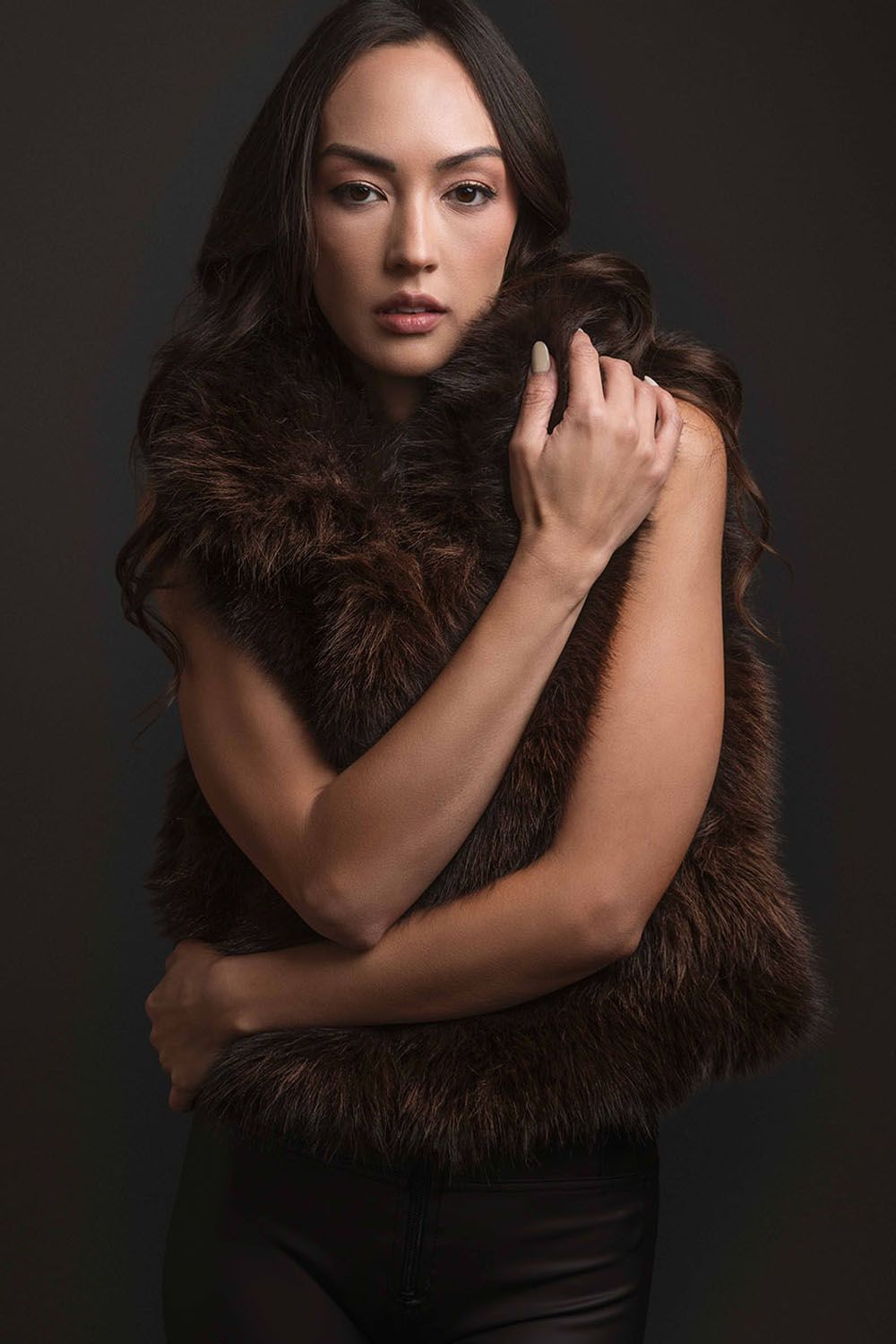 Woman in brown fur vest posing elegantly against dark background with arms crossed.