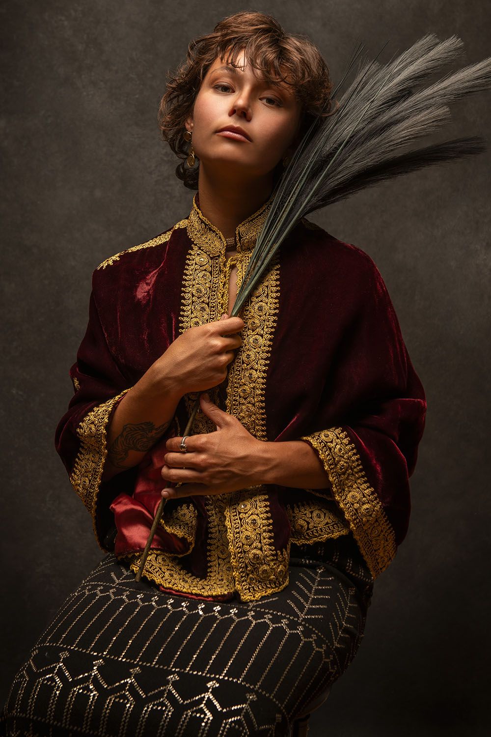 Person in ornate burgundy and gold embroidered jacket holding dried palm leaves, studio portrait on dark background.
