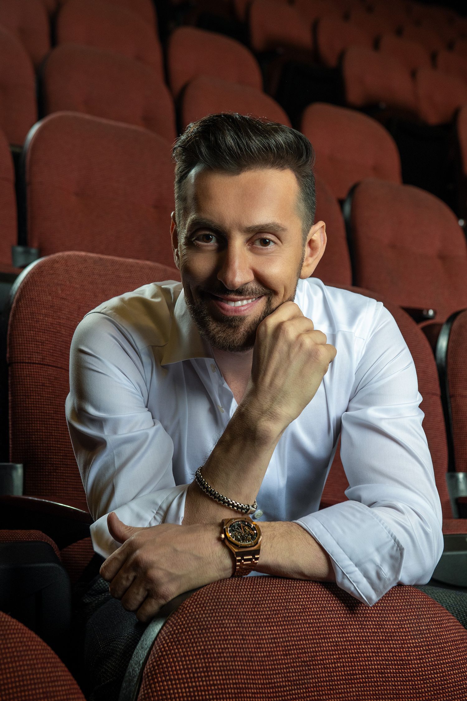 persian Iranian comedian Max Amini in white dress shirt poses thoughtfully in red theater seats during professional photoshoot.