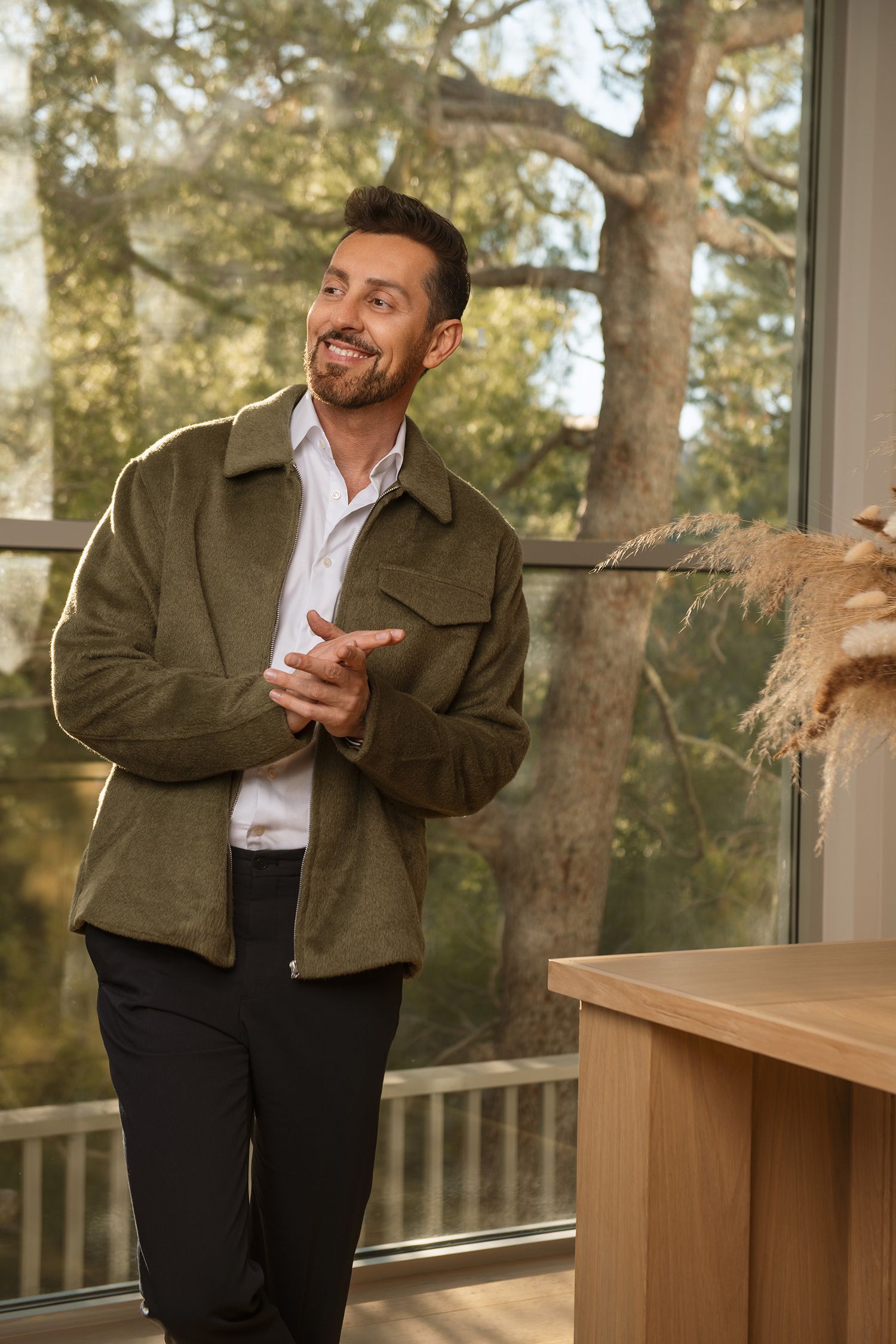 Casual portrait of person laughing and gesturing in olive jacket by sunlit window of comedian Max Amini taken in Westwood Los Angeles studio