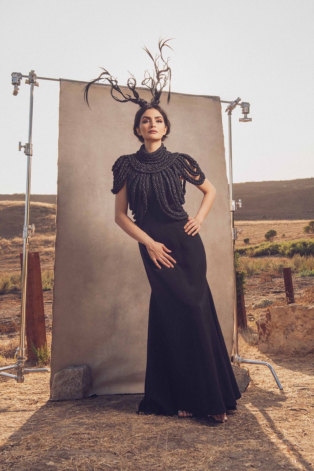 Woman in black gown with beaded top and antler headpiece poses outdoors at a fashion photo shoot.