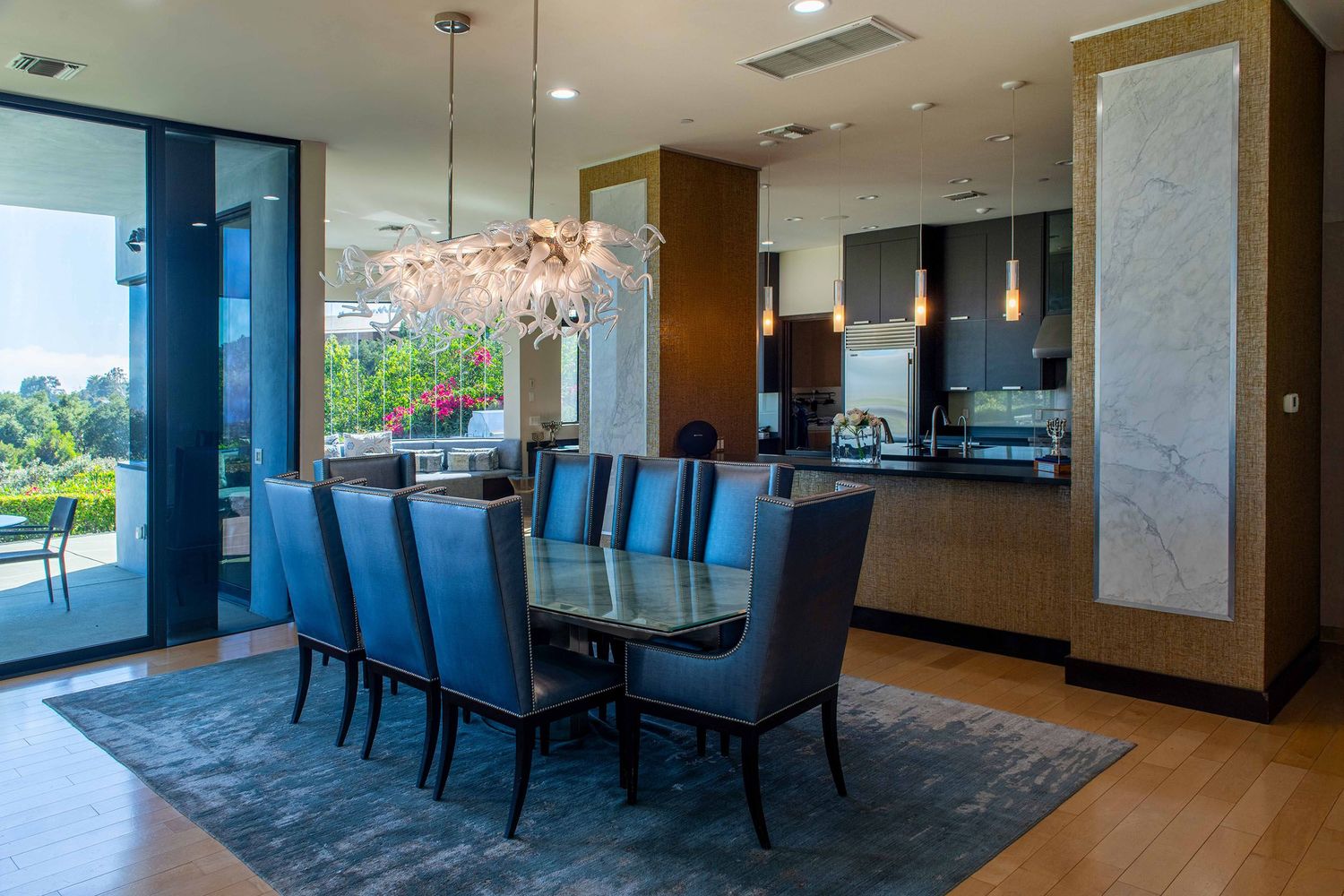 Modern dining room with blue upholstered chairs, marble walls, and a contemporary glass chandelier overlooking outdoor views.