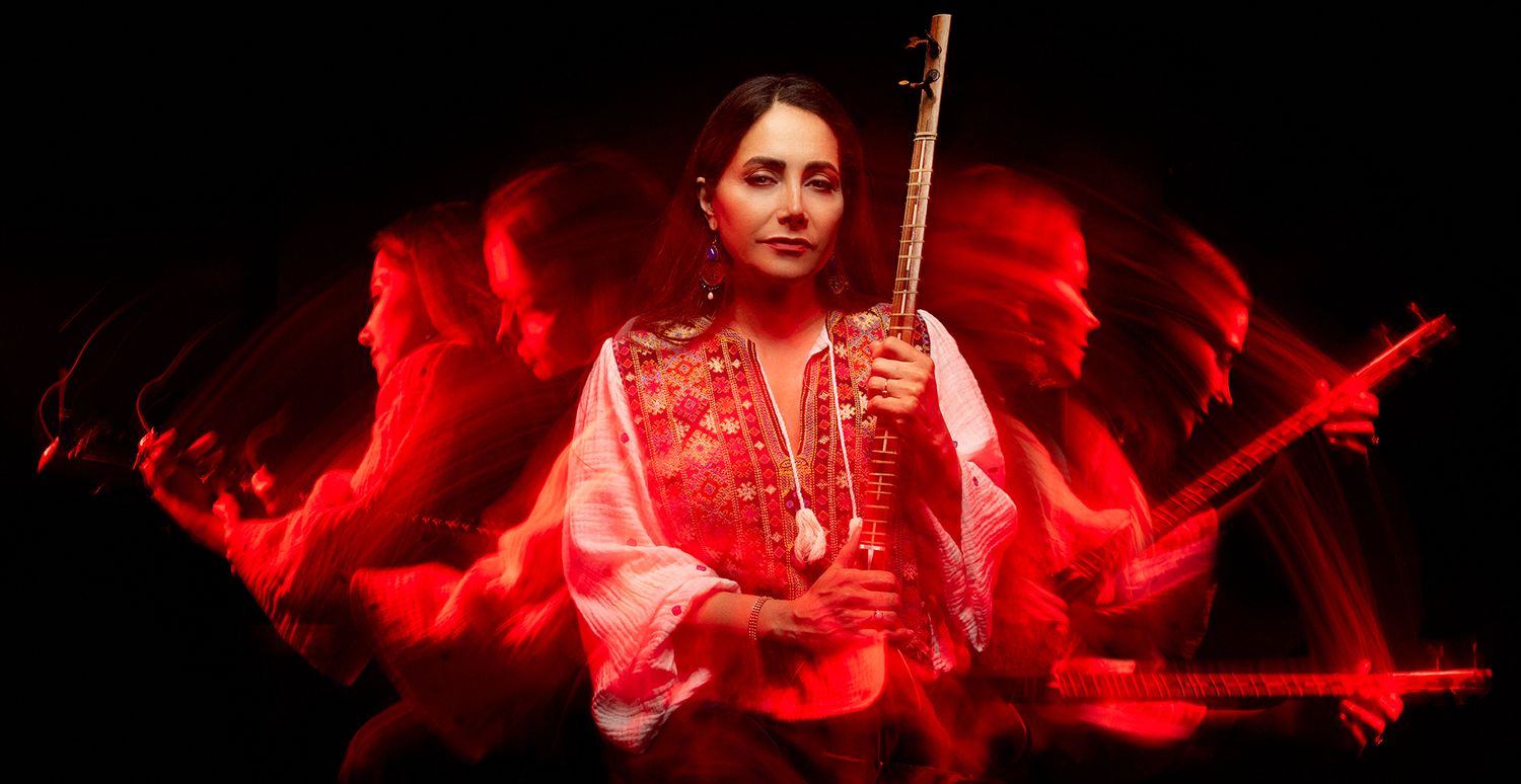 Woman in pink embroidered top playing a stringed instrument against dramatic red light background.