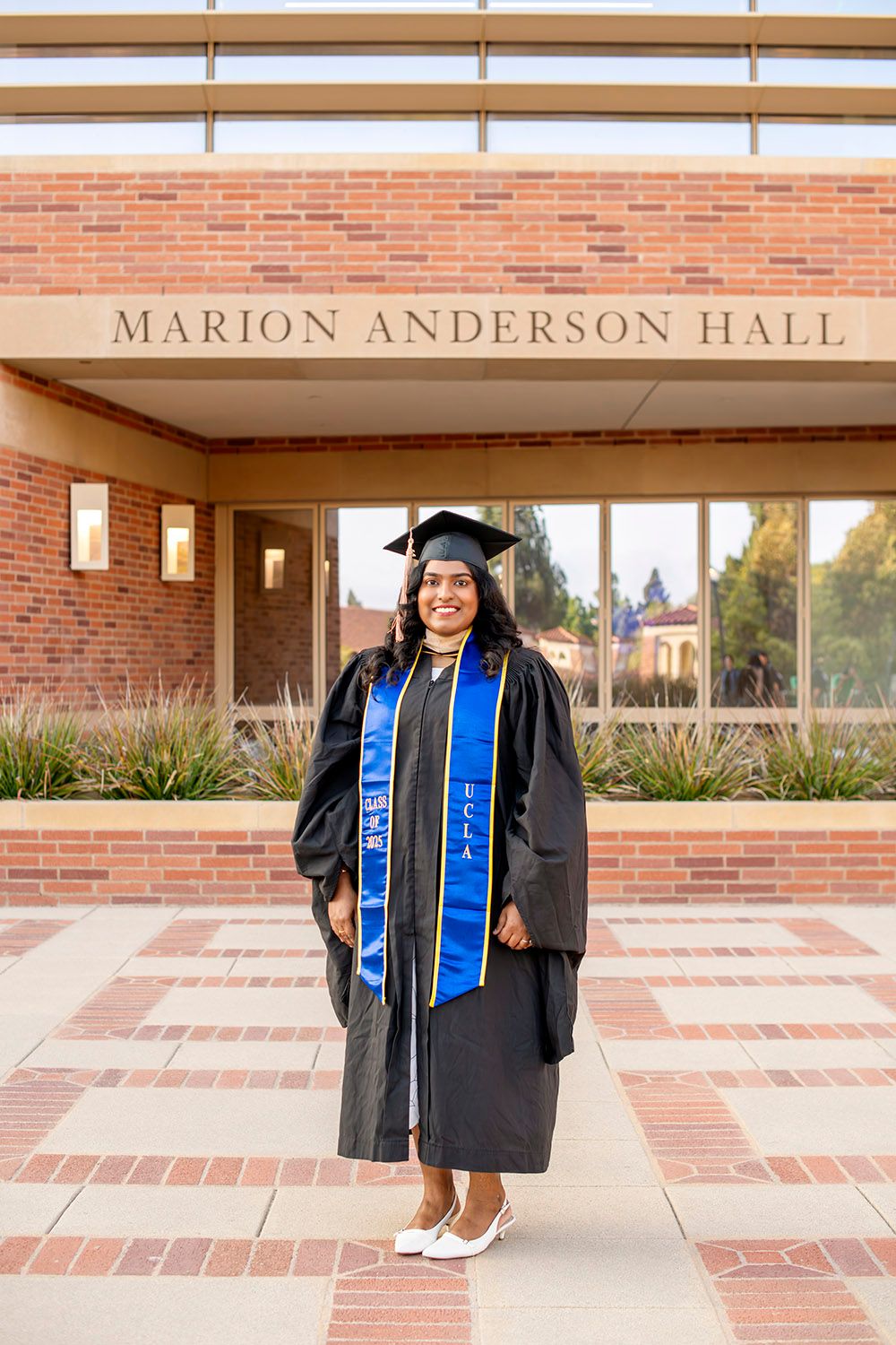 Graduate in cap and gown with blue UCLA stole poses proudly in front of Marion Anderson Hall on campus.
