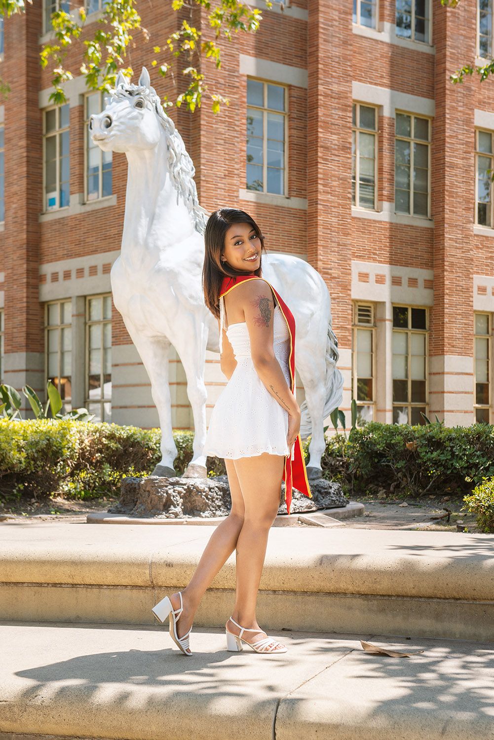 Young woman in white dress poses beside a white horse statue outside a red brick university building on a sunny day.