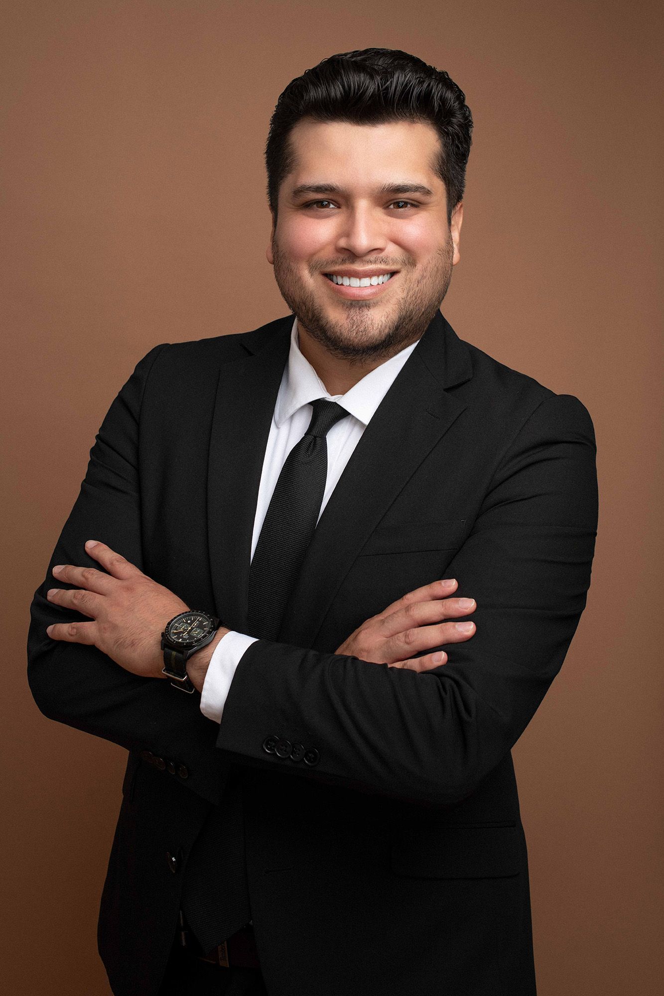 Smiling businessman in black suit and tie with arms crossed against brown background.