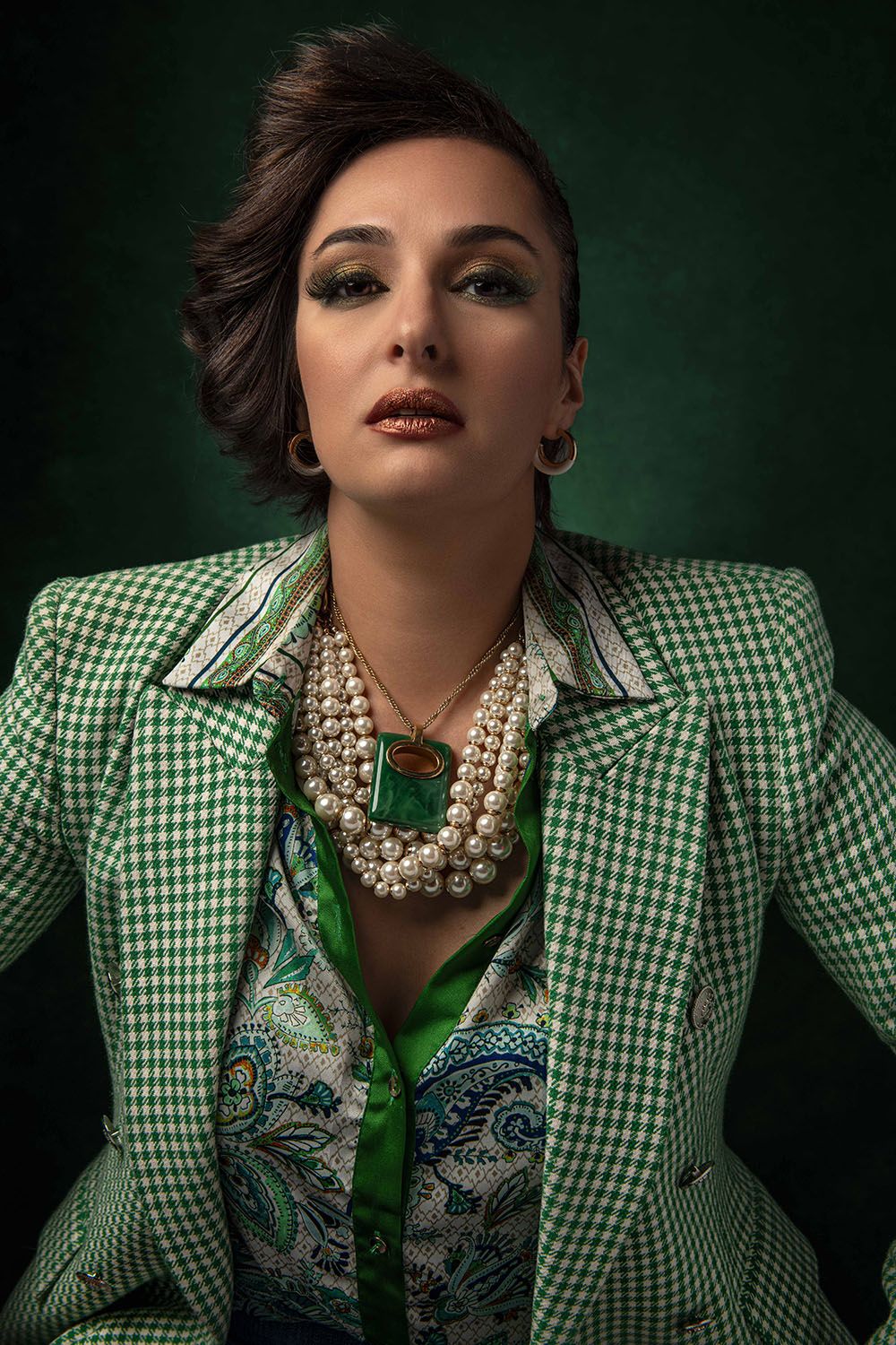 Woman in green houndstooth blazer with pearl necklace and paisley blouse poses against dark green background.
