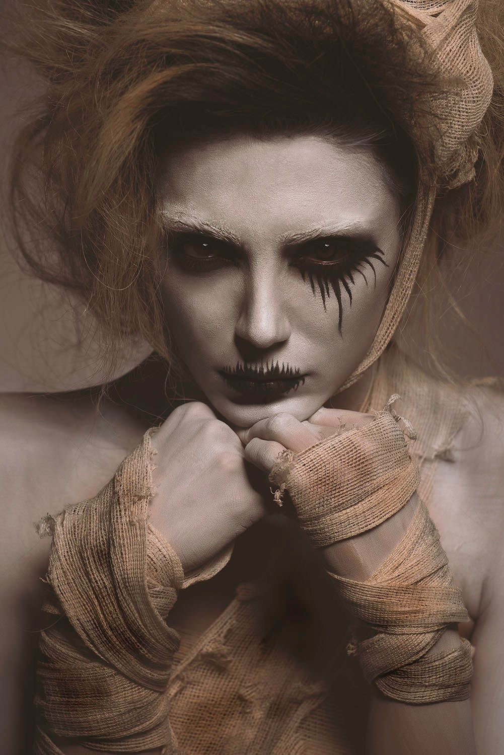 Woman with dramatic Halloween mummy makeup and dark eye paint with bandage-wrapped hands poses for eerie portrait.