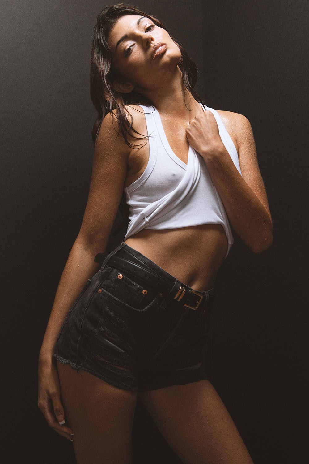 Woman in white crop top and denim shorts posing against dark background.