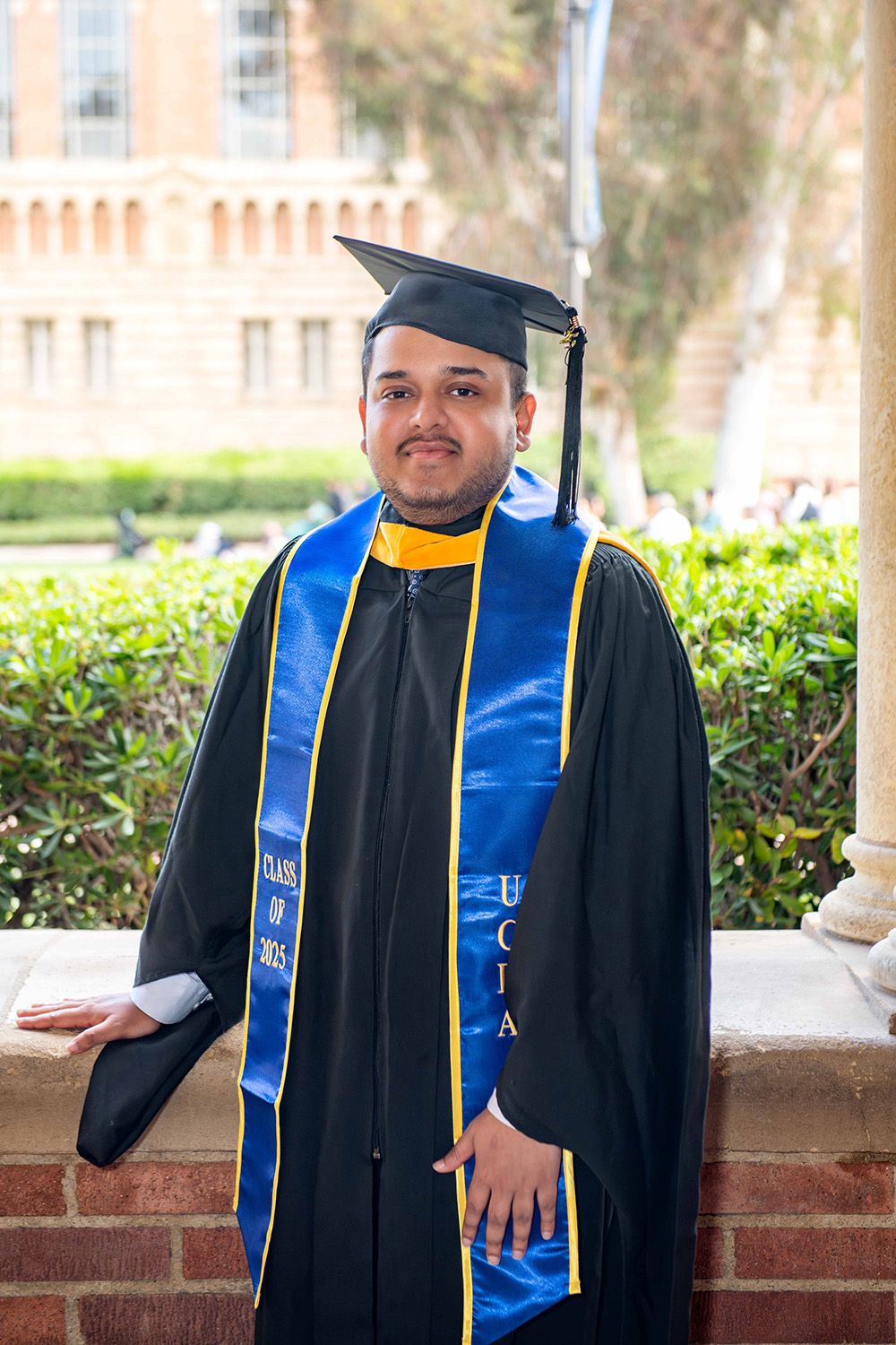 UCLA graduate smiles in black cap and gown with blue and gold stole outdoors on campus.
