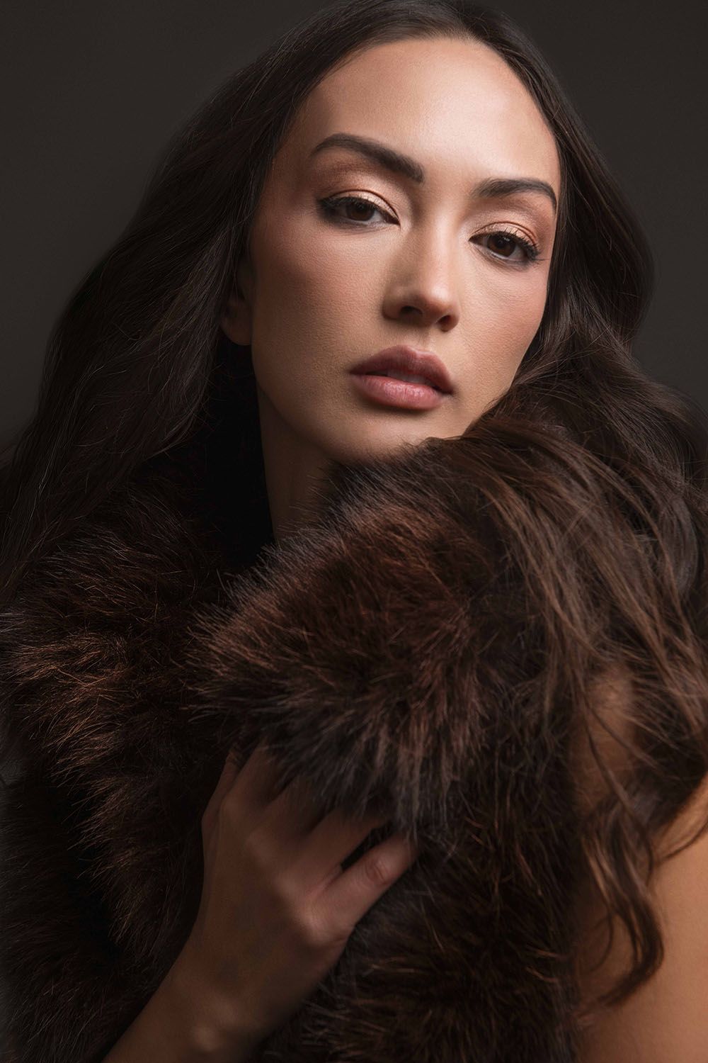 A woman with long dark hair poses elegantly wrapped in a luxurious brown fur stole against a dark background.