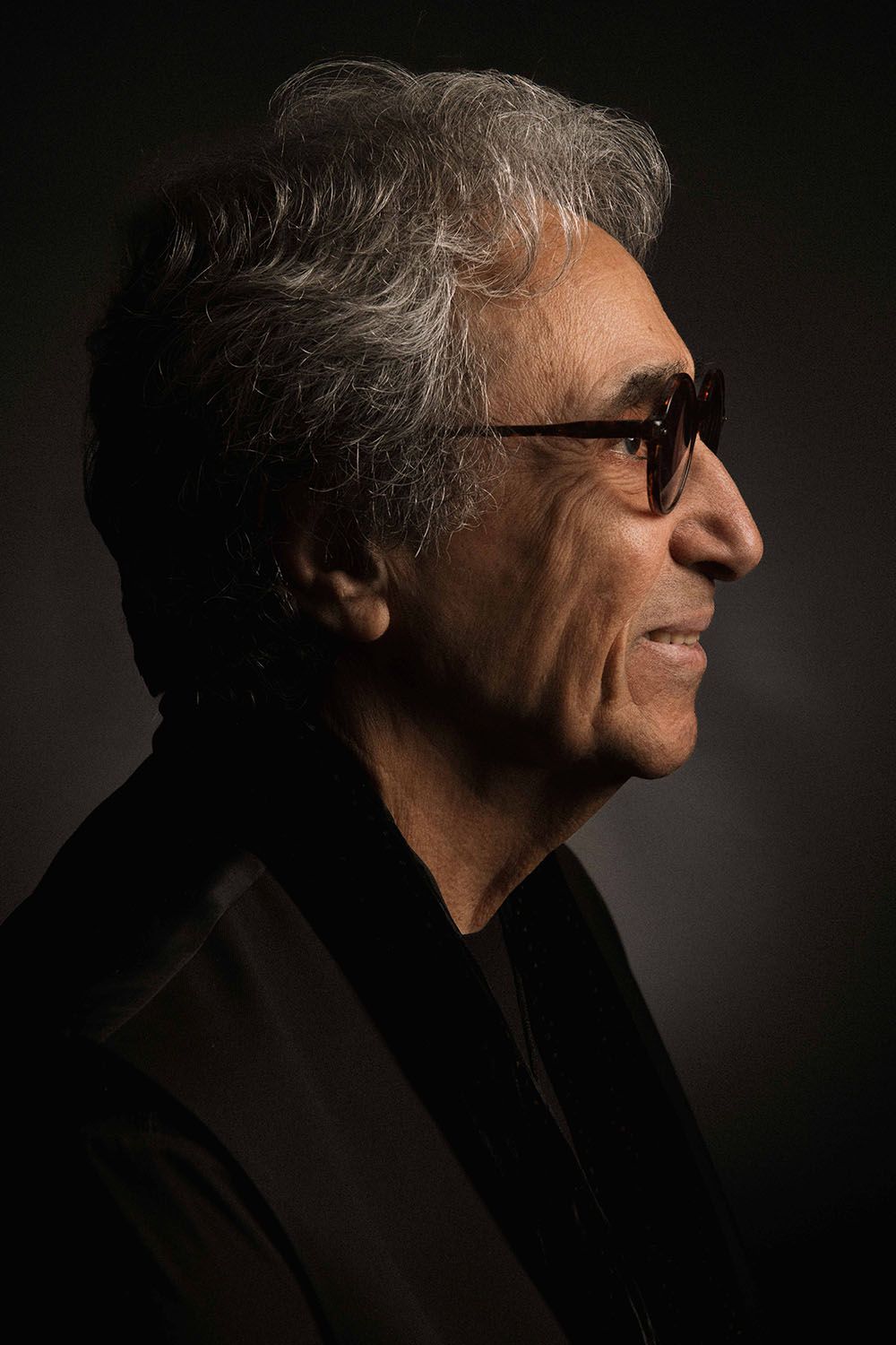 Side profile of an older man with gray hair wearing dark sunglasses and a black blazer against a dark background.