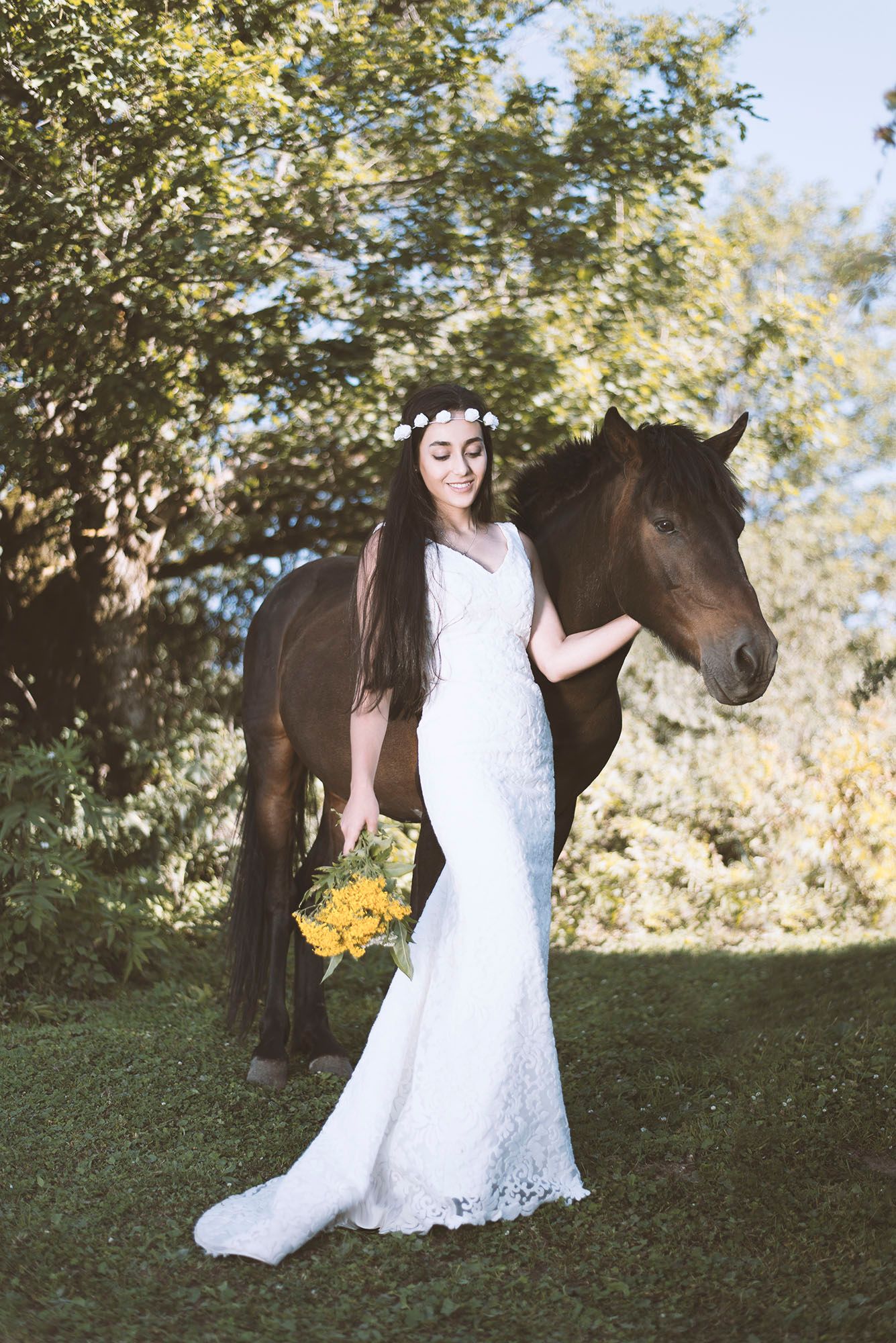 A bride in a white lace dress poses with a dark brown horse in a grassy outdoor setting with yellow sunflowers.