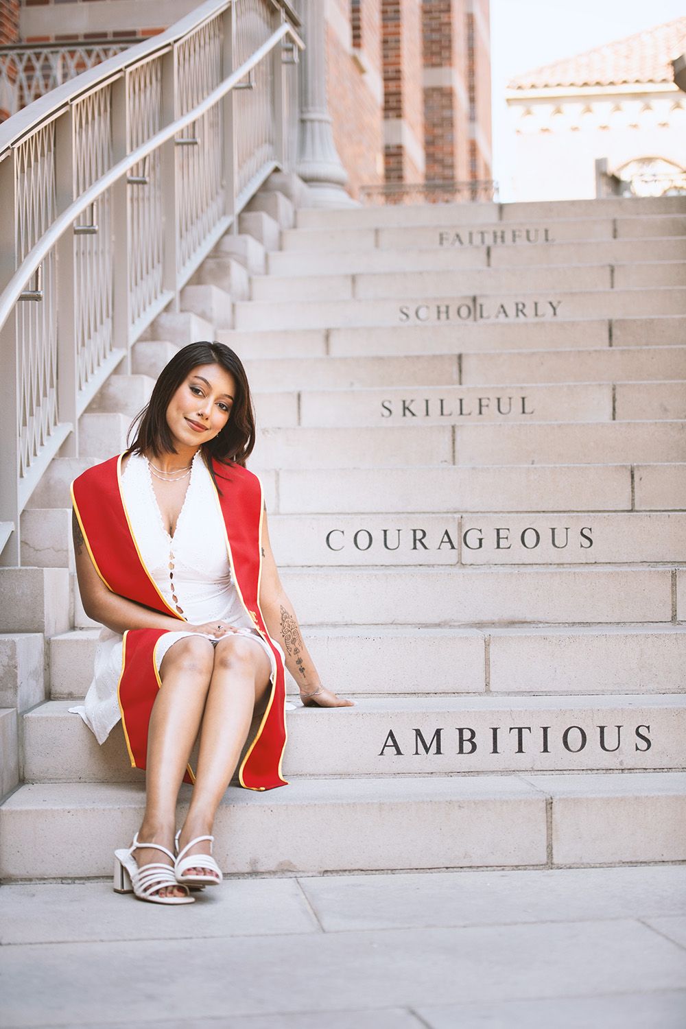 Graduate woman sitting on university steps wearing red academic regalia, surrounded by inspirational words like Ambitious.