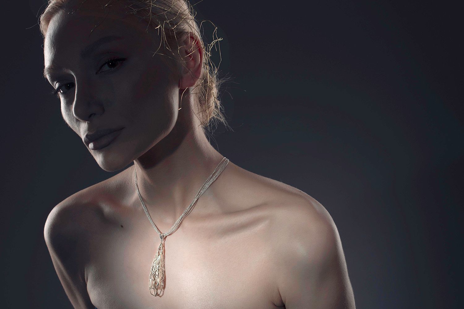 Elegant woman wearing a delicate gold pendant necklace against a dark background in a dramatic studio portrait.