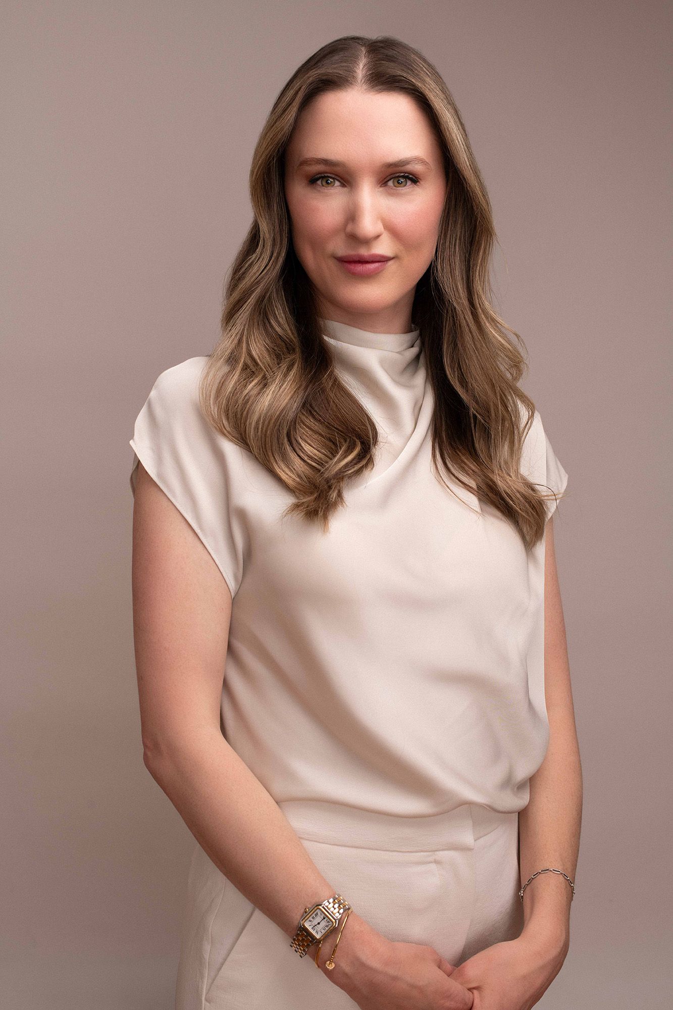Professional woman in white sleeveless mock-neck top with gold watch, posing against neutral beige background.