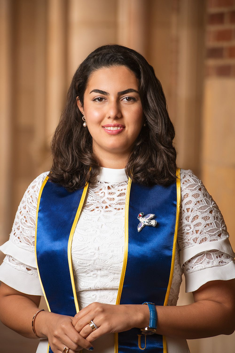 Young woman in white lace dress wearing blue and gold graduation stole smiles for professional portrait photo.