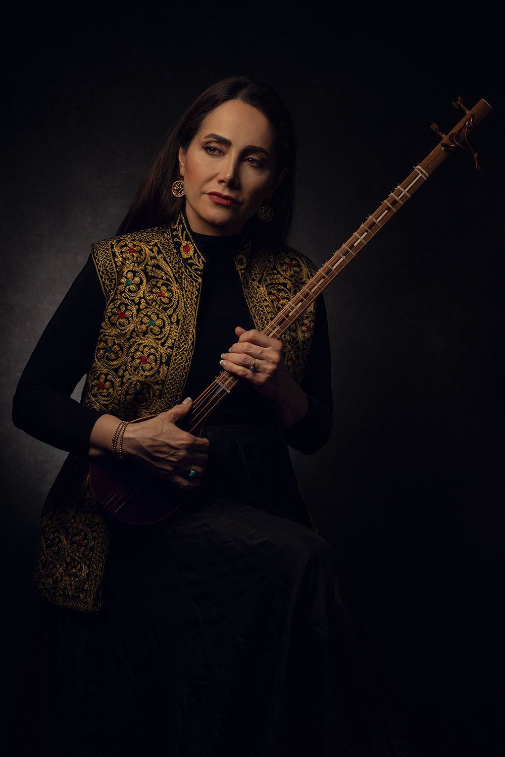 Woman in embroidered vest holding a traditional setar instrument against a dark background.