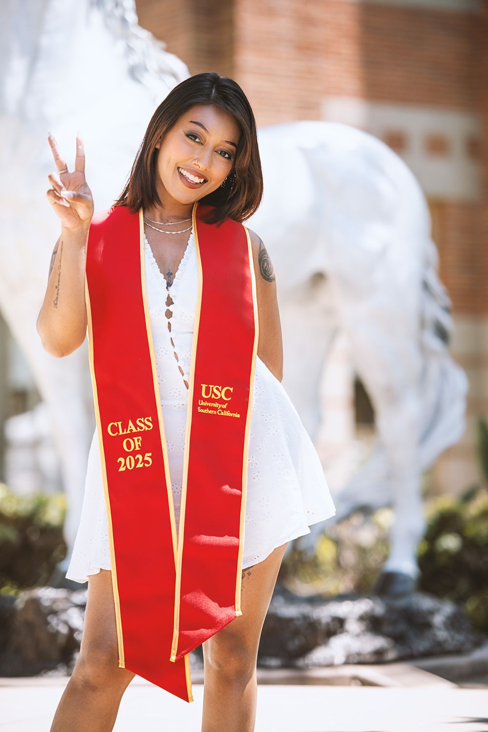 USC Class of 2025 graduate in red stole poses with peace sign in front of white Trojan horse statue.