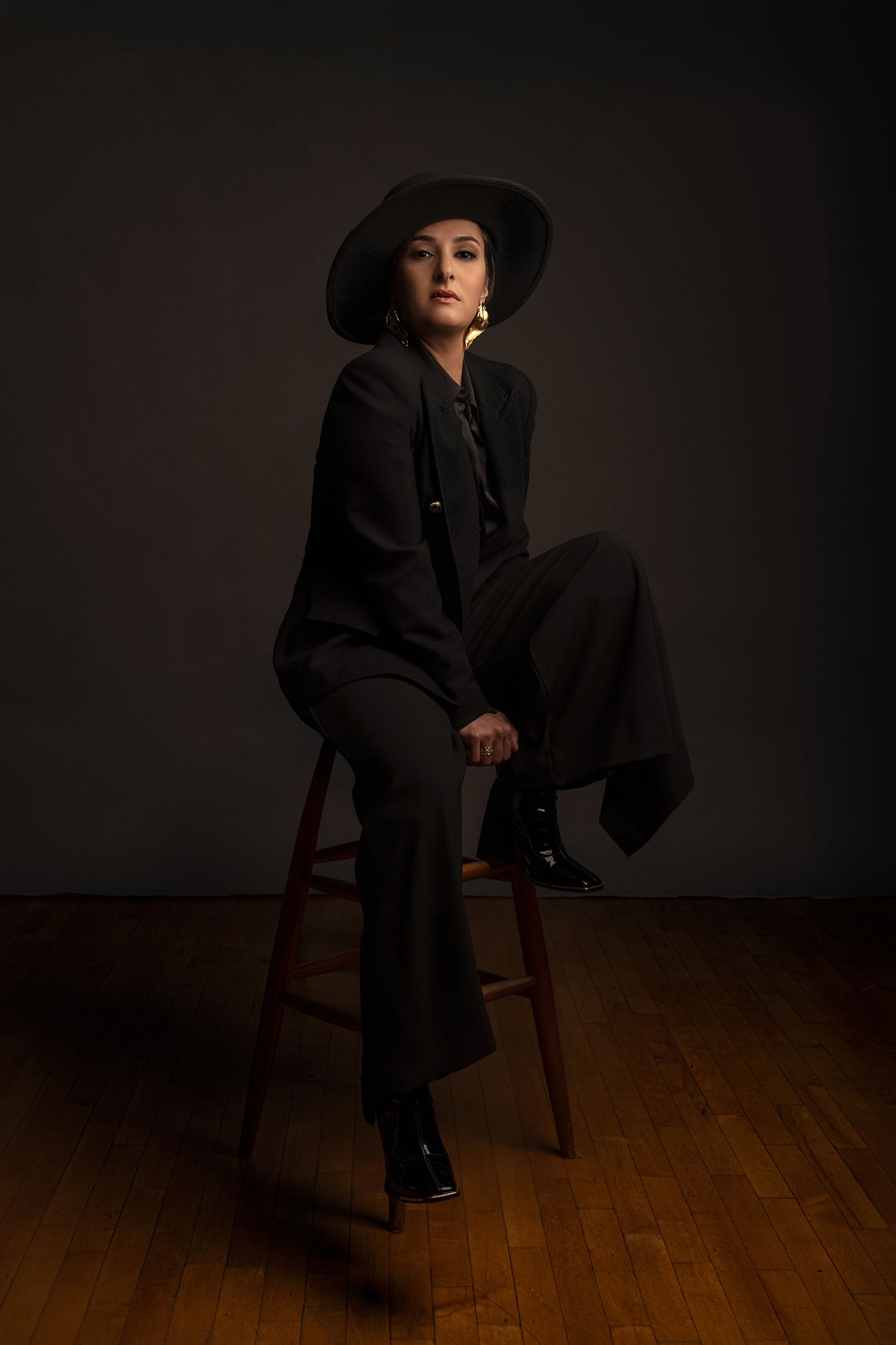 Dramatic portrait of persian pop singer Rana Mansour featuring figure in black suit and hat seated on wooden chair against dark backdrop.
