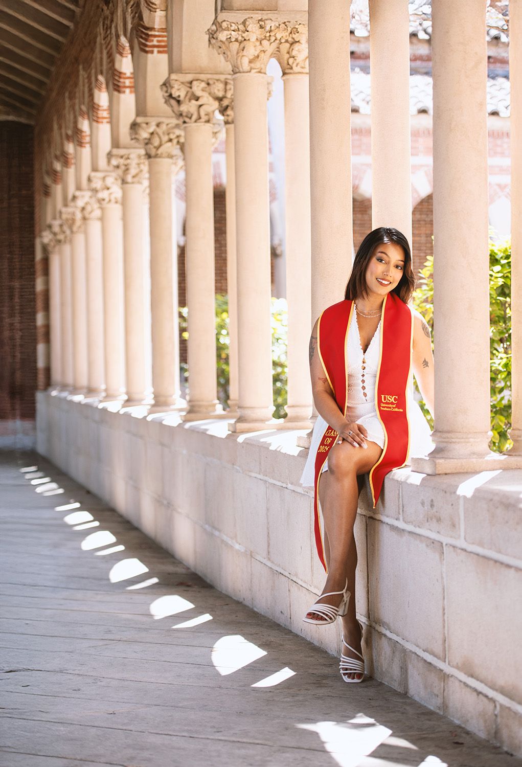 A young woman in a white dress with a red USC graduation sash poses along a sunlit columned walkway.