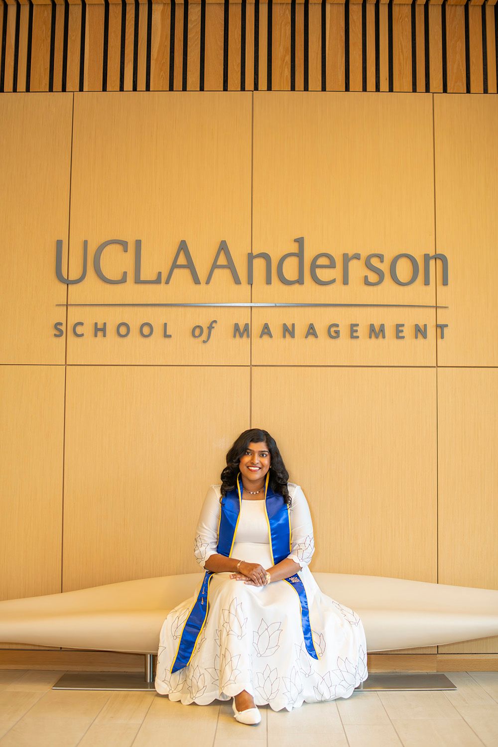 UCLA Anderson School of Management graduate smiles in white dress with blue sash, seated in front of the iconic school sign.