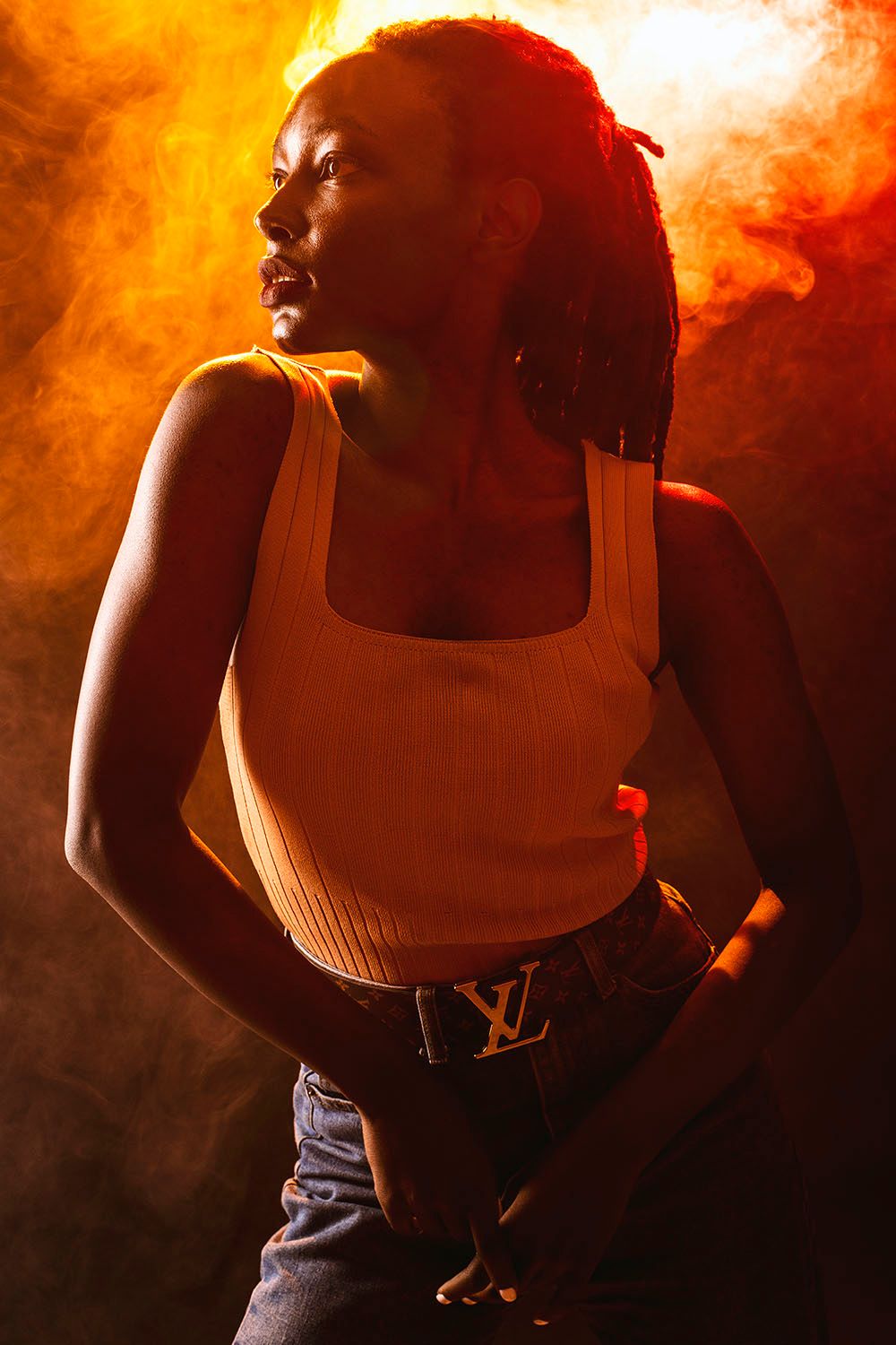 Young Black woman with dreadlocks wearing a white tank top and Louis Vuitton belt poses against dramatic orange smoke background.