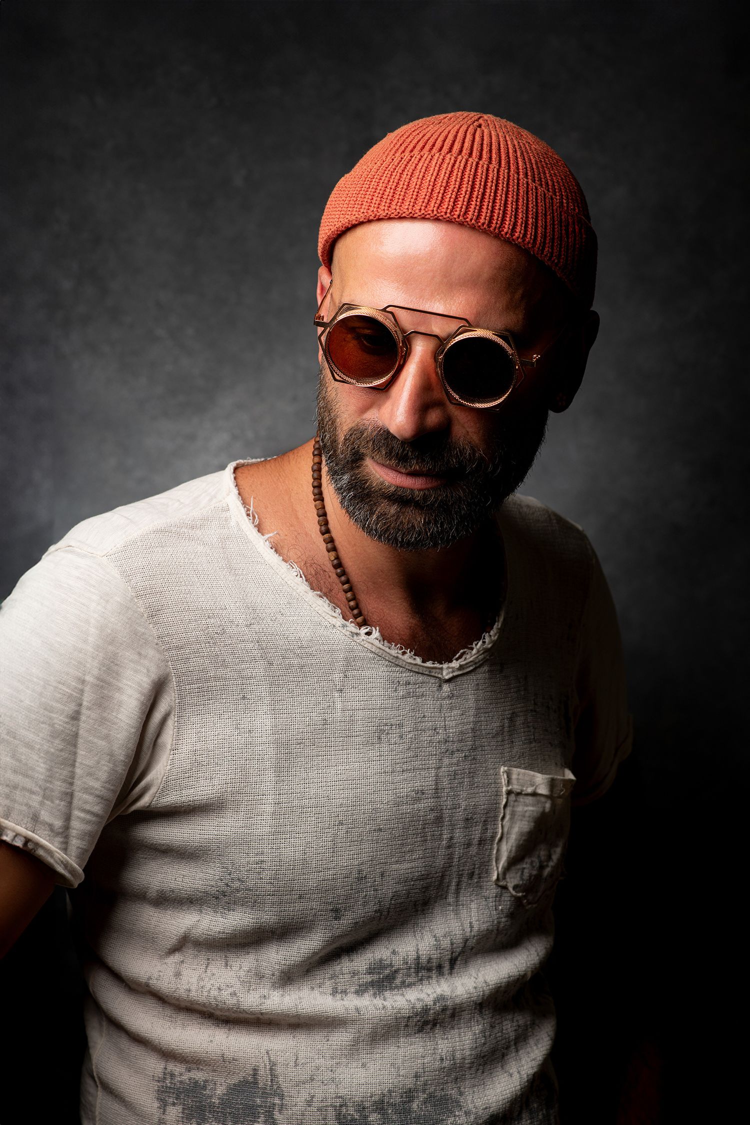 persian podcaster and comedian Farshid Manafi wearing orange beanie and sunglasses poses against dark backdrop in casual attire.