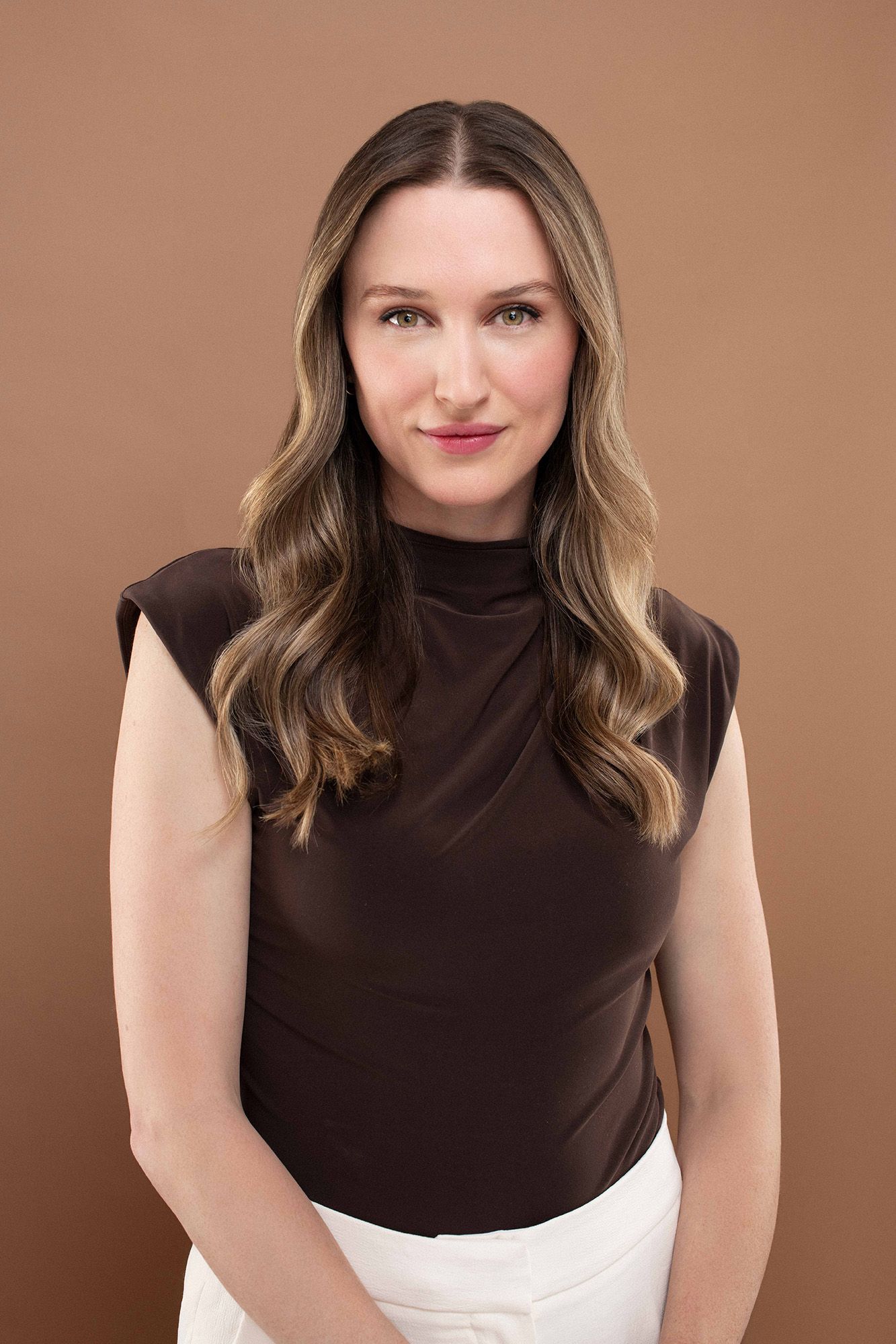 Woman in brown sleeveless turtleneck top posing confidently against a warm brown studio background.