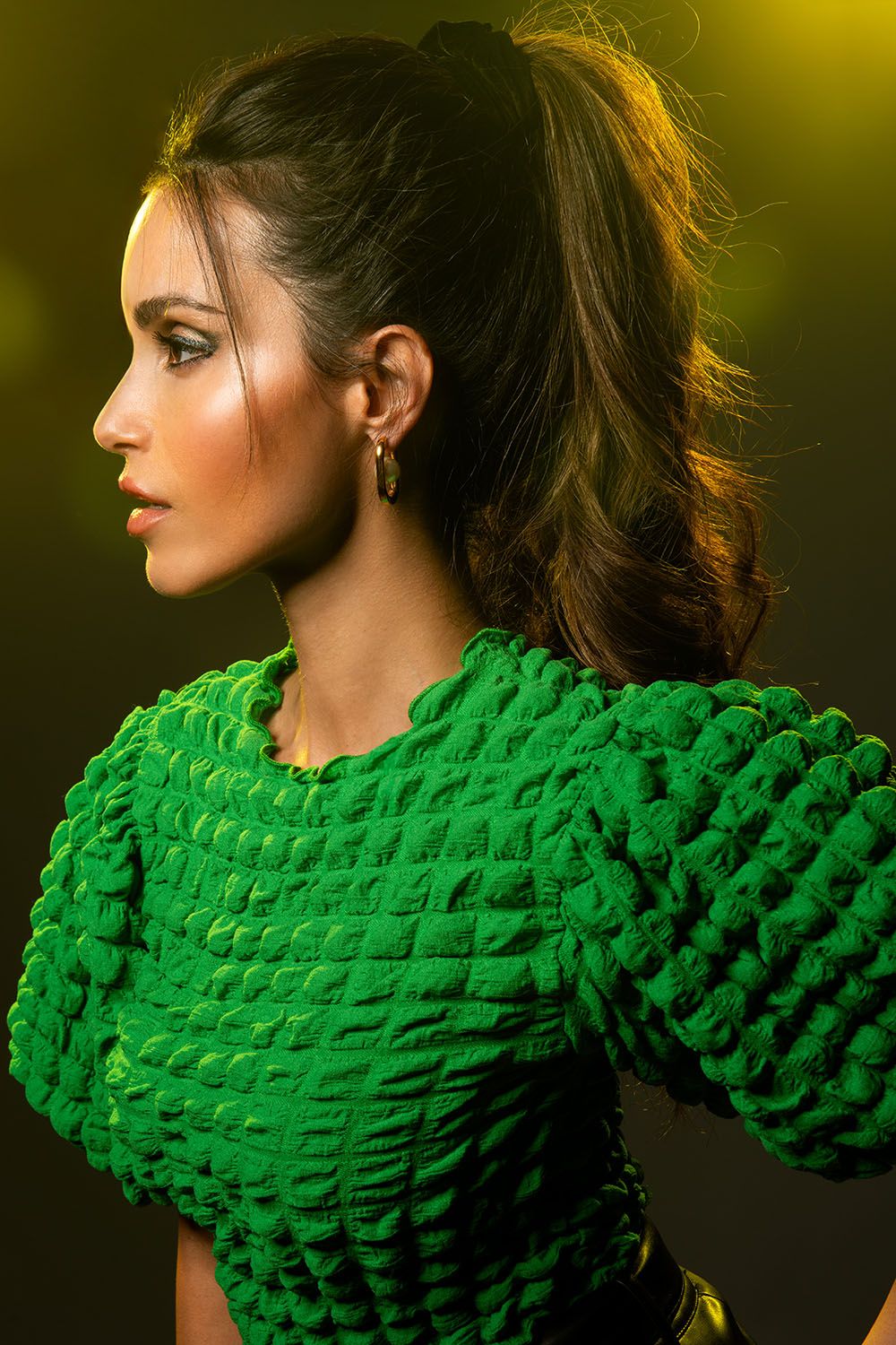 Woman in textured green dress with ponytail poses in profile against golden yellow background.