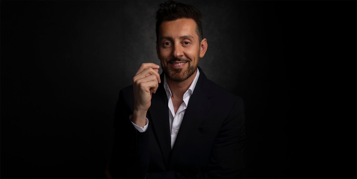 Professional studio portrait of a person in a black suit jacket and white shirt against a dark background.