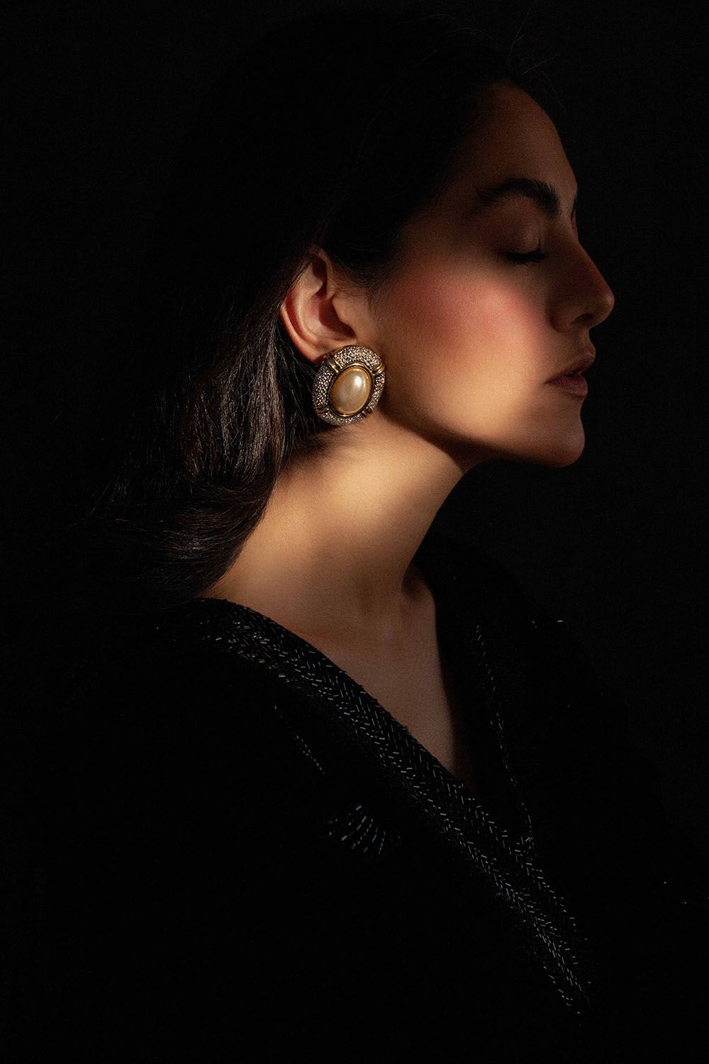 Woman in profile with closed eyes wearing large ornate pearl and crystal earring against dark background.
