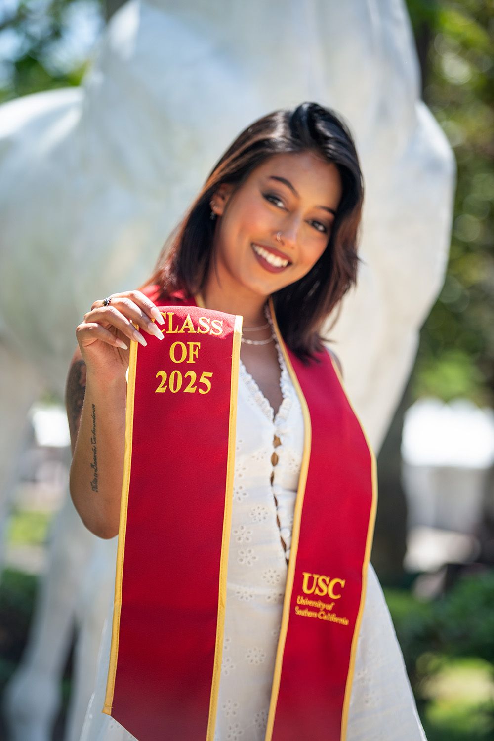 A smiling USC Class of 2025 graduate poses in a white dress wearing a red and gold USC graduation stole outdoors.