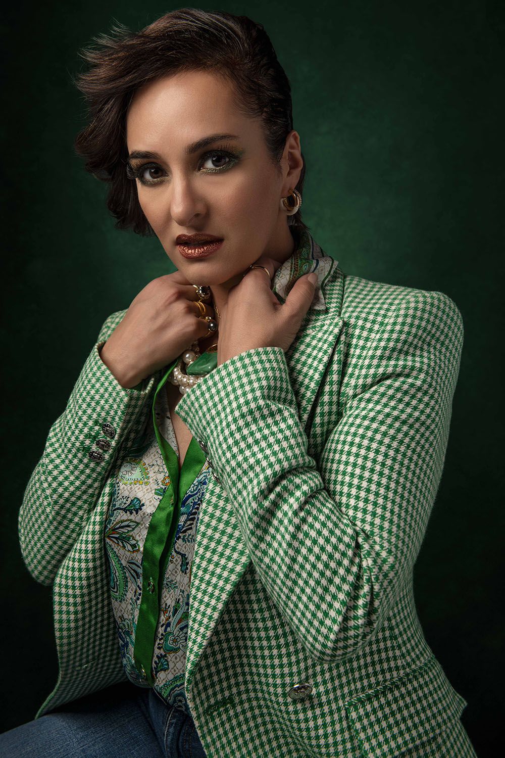 Woman in green checkered blazer posing confidently with hands near neck, wearing gold jewelry on dark green background.