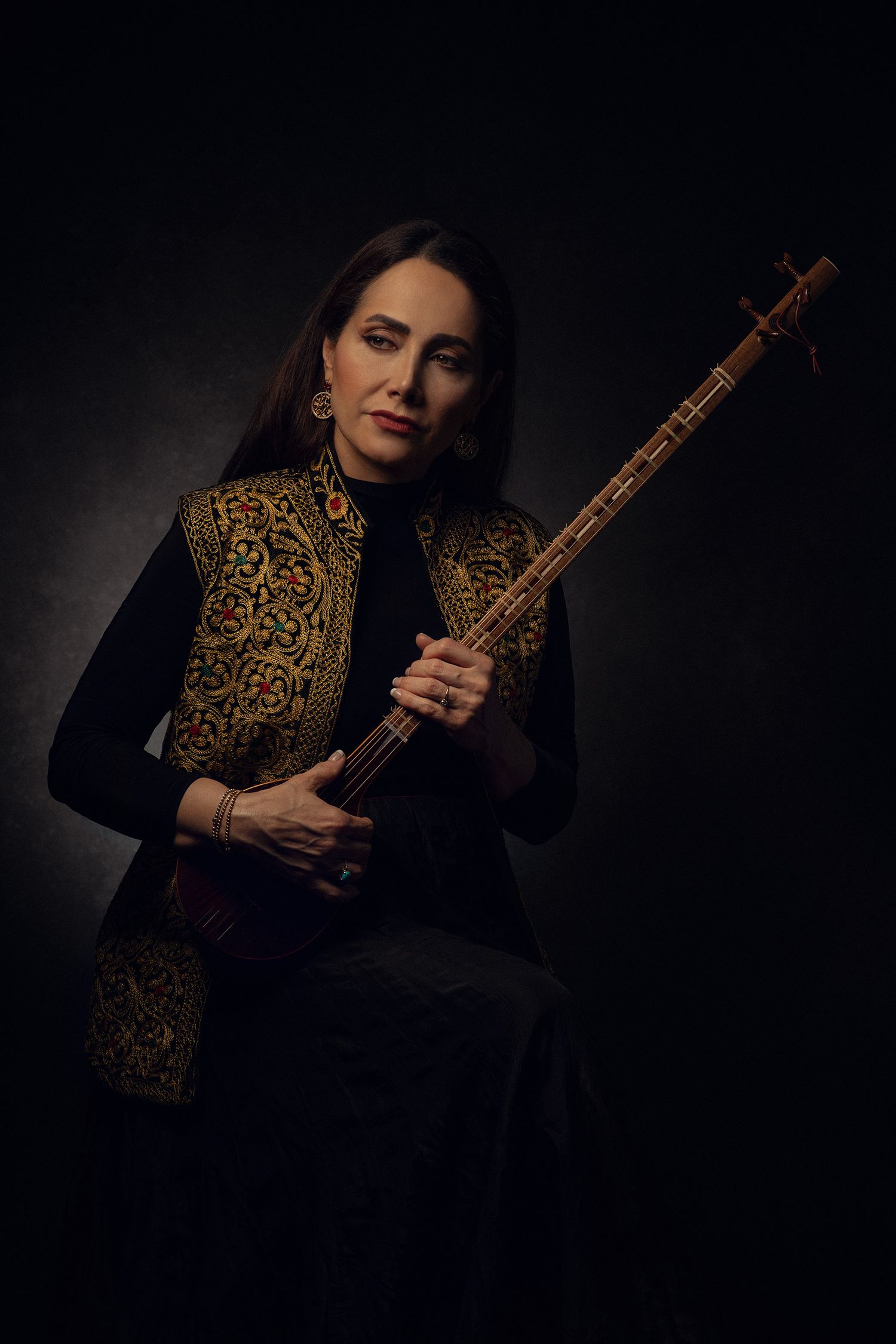Classical musician and Iranian singer Mojgan Shajarian in ornate vest holds traditional sitar instrument against dark background.